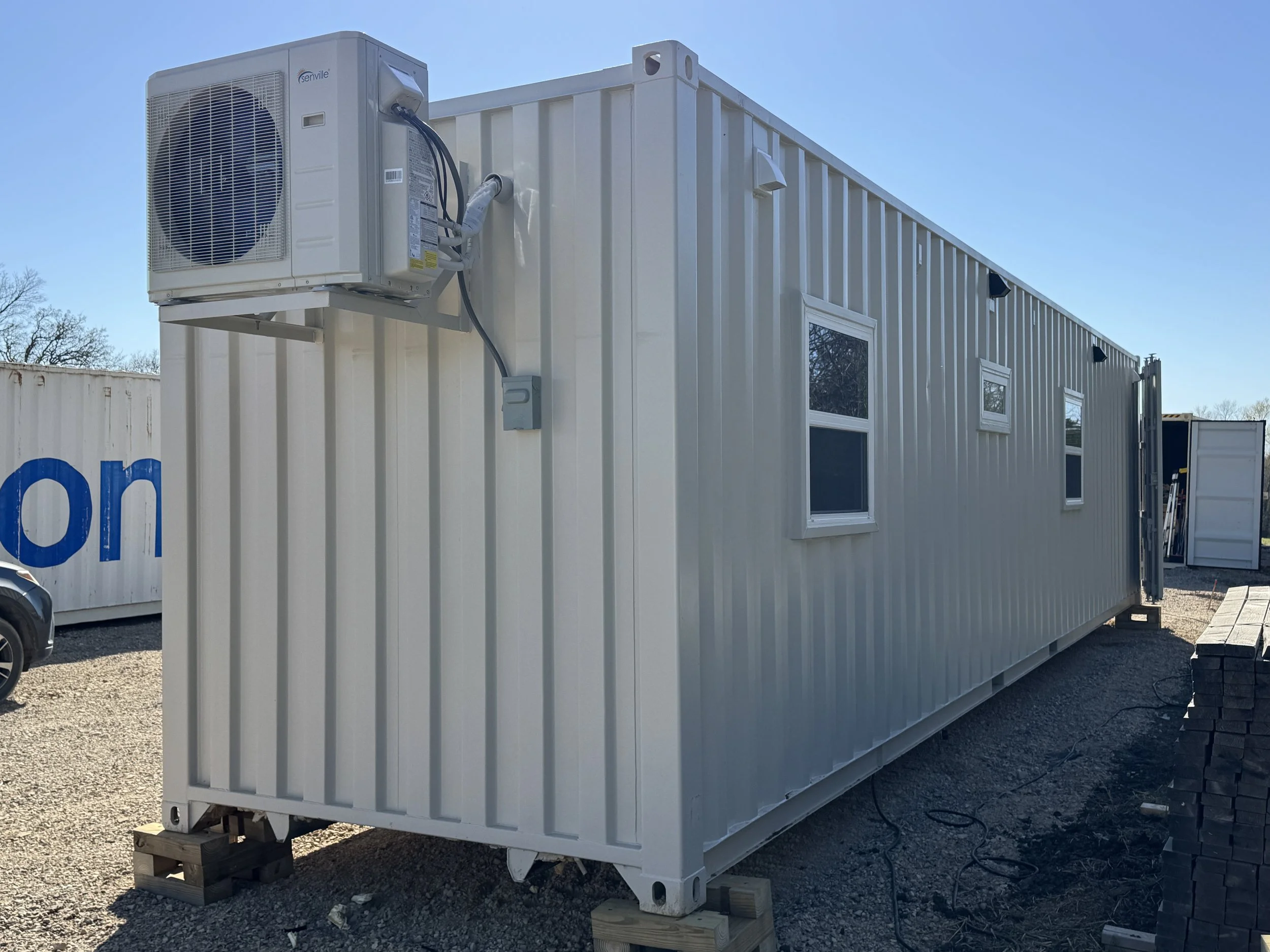 Turnkey shipping container home side exterior in Paris Texas with wall-mounted Daikin mini-split air conditioner, white steel siding, windows, and gravel lot under clear blue sky