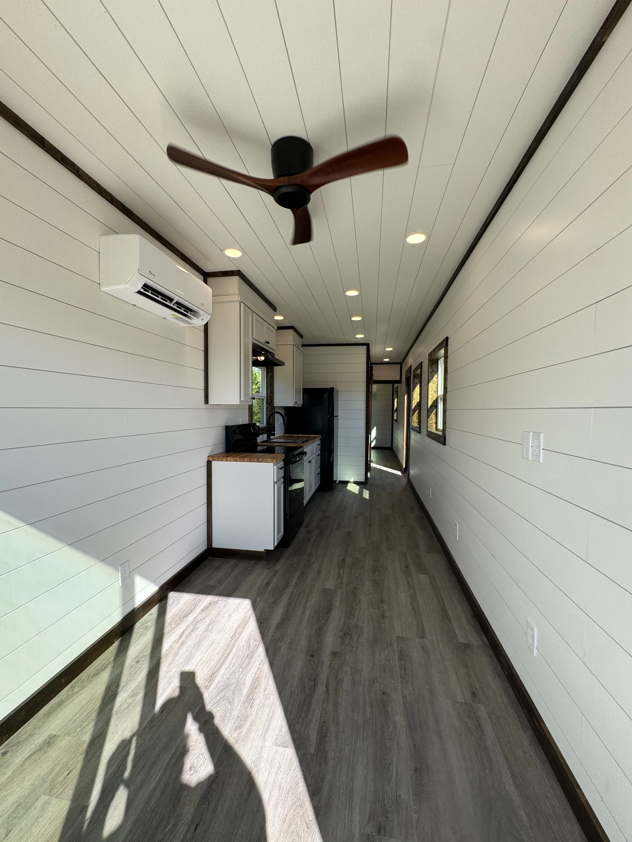 Long interior hallway view inside turnkey shipping container home in Paris Texas with white shiplap walls, kitchen area featuring white cabinets and black appliances, wall-mounted mini-split AC unit, wood-look flooring, and natural light from windows