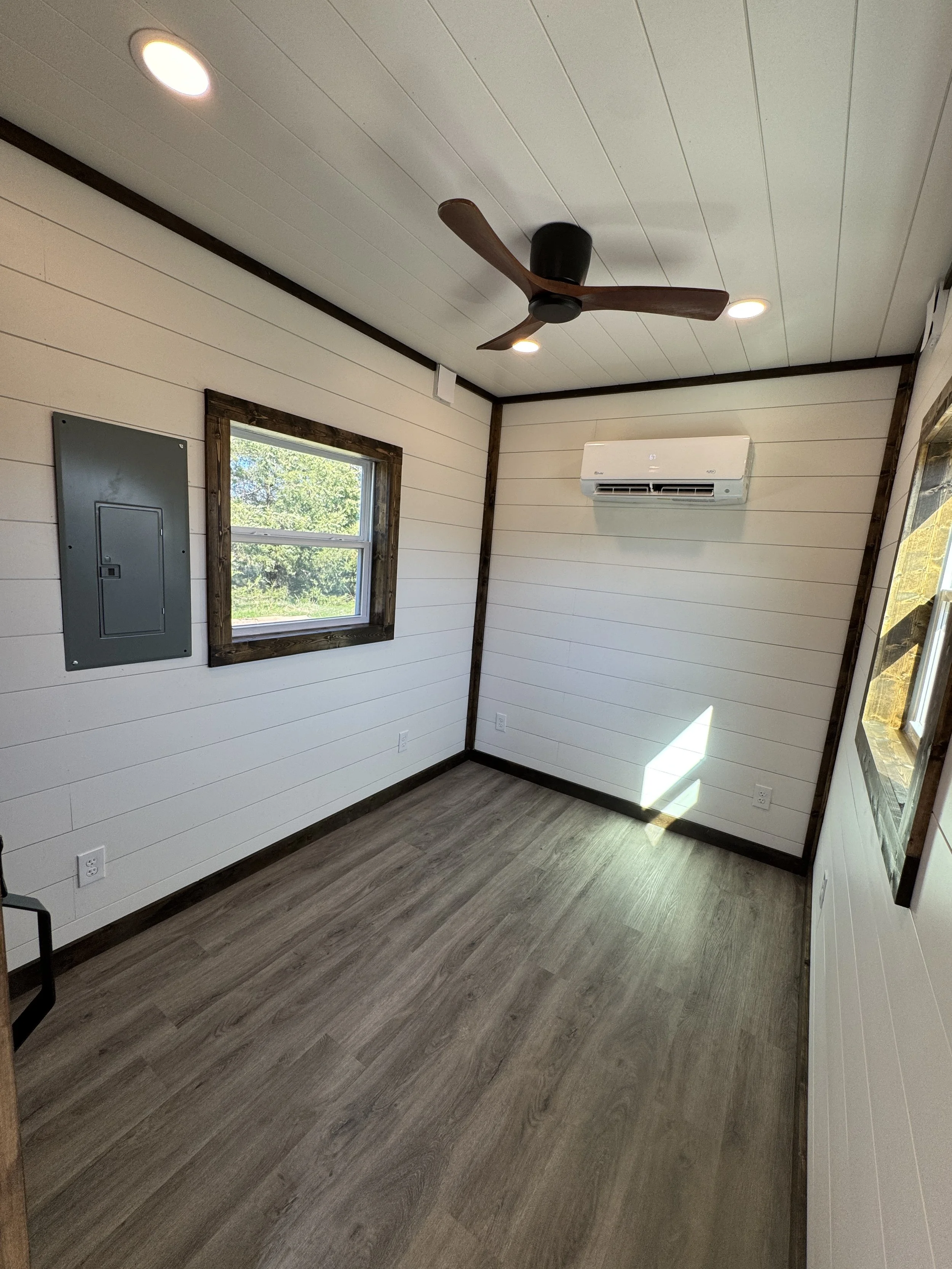Turnkey shipping container home interior in Paris Texas with white shiplap walls, dark wood ceiling fan, large window, wall-mounted mini-split air conditioner, electrical panel, and wood-look flooring