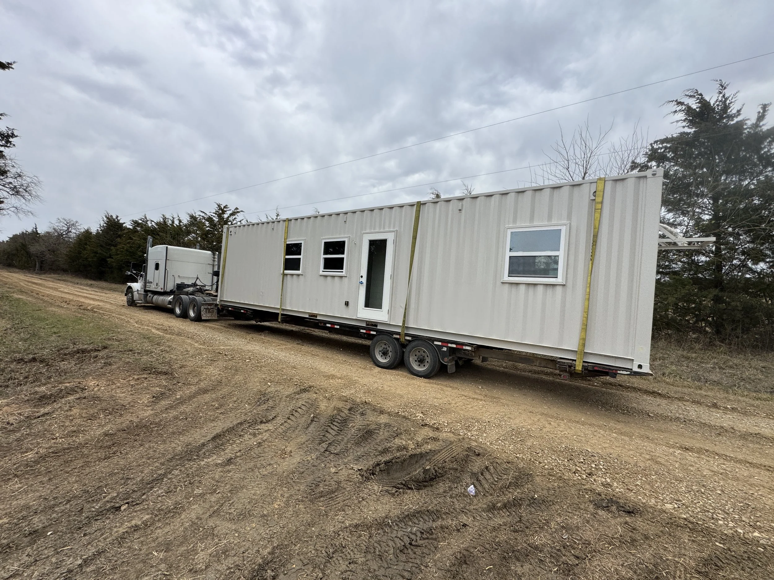 Professional delivery of turnkey shipping container home in Paris Texas on a flatbed trailer with semi truck on gravel road under cloudy sky