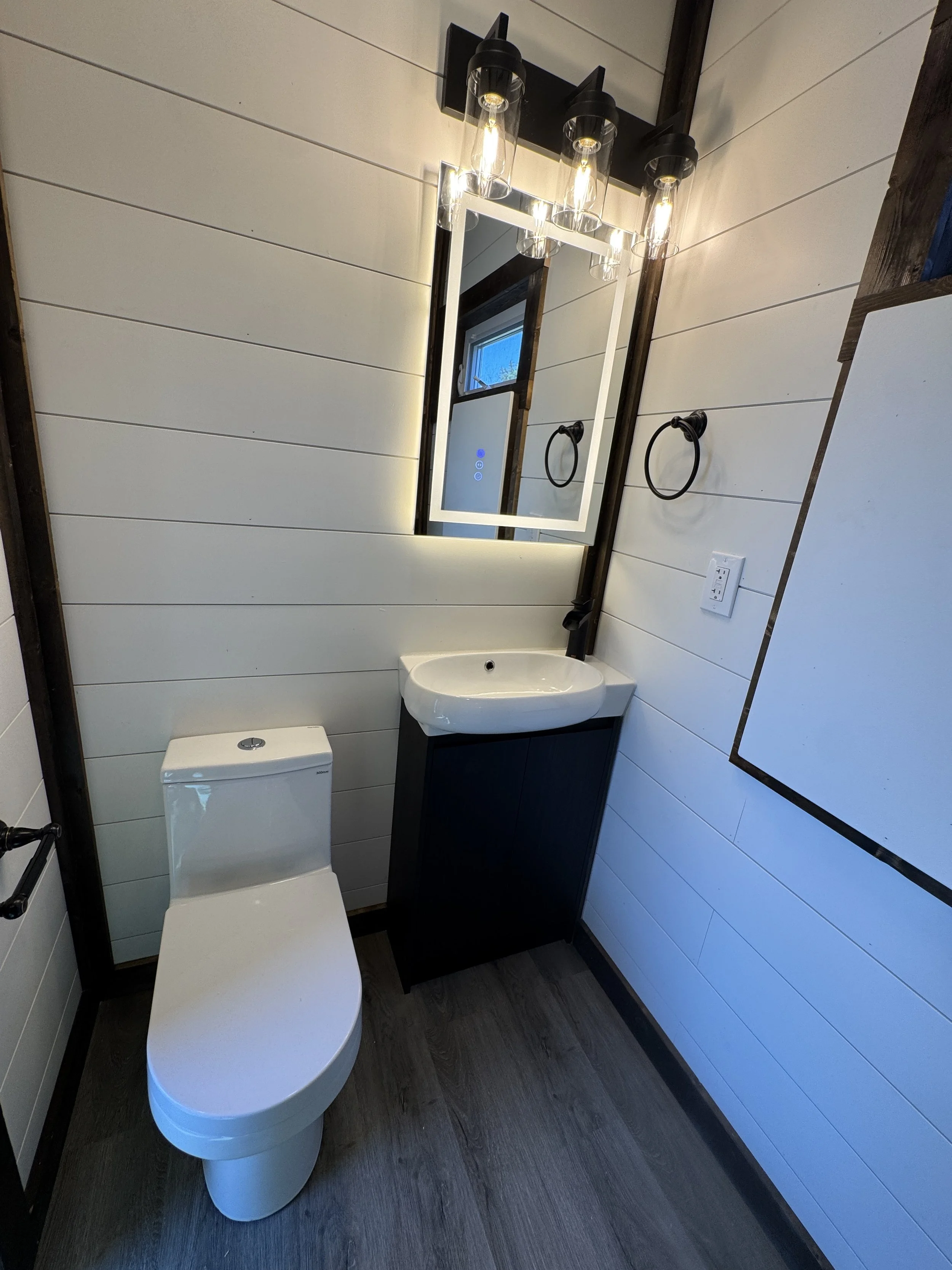 Modern bathroom interior in turnkey shipping container home in Paris Texas with white toilet, black vanity, white vessel sink, large backlit mirror with vanity lighting, and white shiplap walls