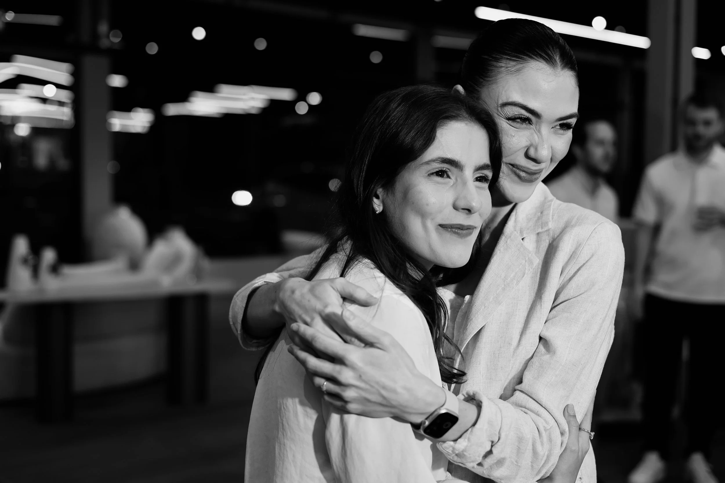 Duas mulheres se abraçando e sorrindo, em um ambiente com fundo com pessoas ao redor.