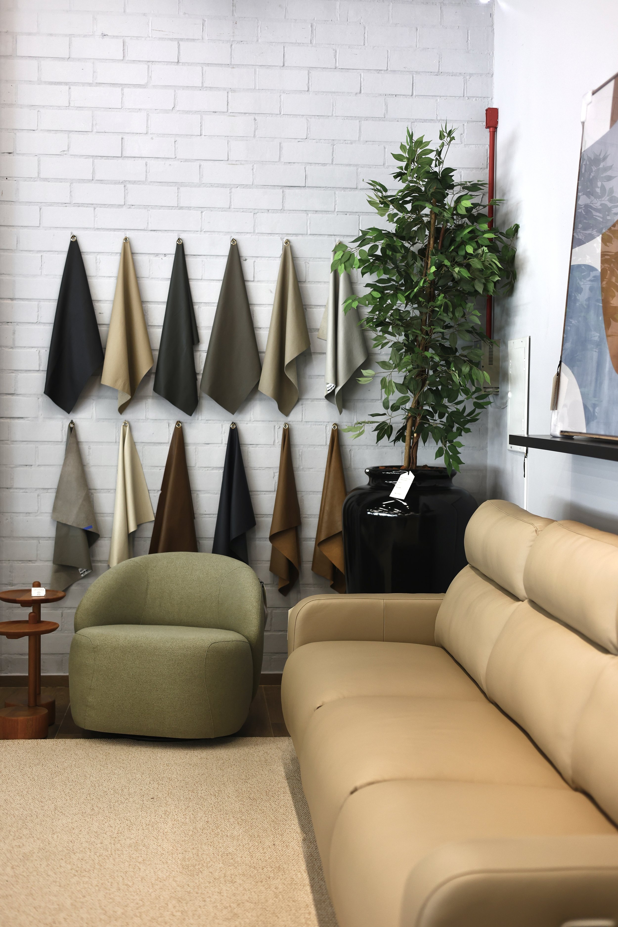 Sala de estar com parede de tijolos brancos, exibição de amostras de tecidos coloridos pendurados na parede, sofá bege, cadeira verde, tapete bege, uma planta grande em um vaso preto e uma pequena mesa de centro de madeira.