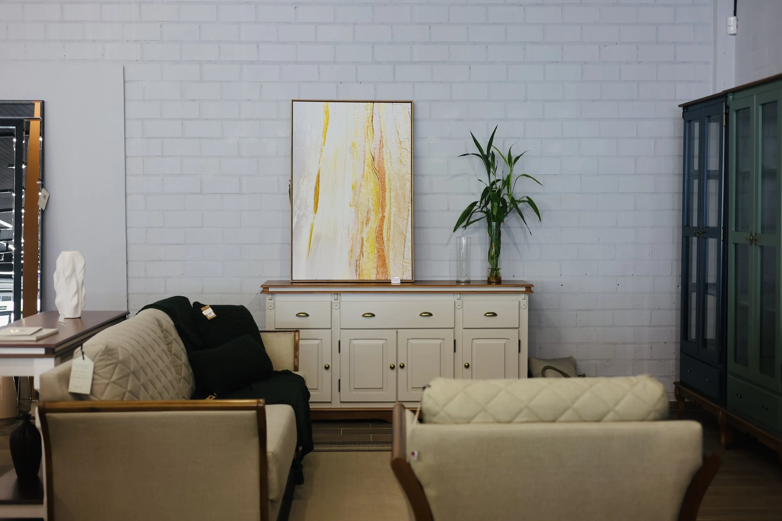 Interior de uma loja de móveis com sofá, quadros, uma estante azul escura, e uma cômoda branca com uma planta ao lado, parede de tijolos brancos.