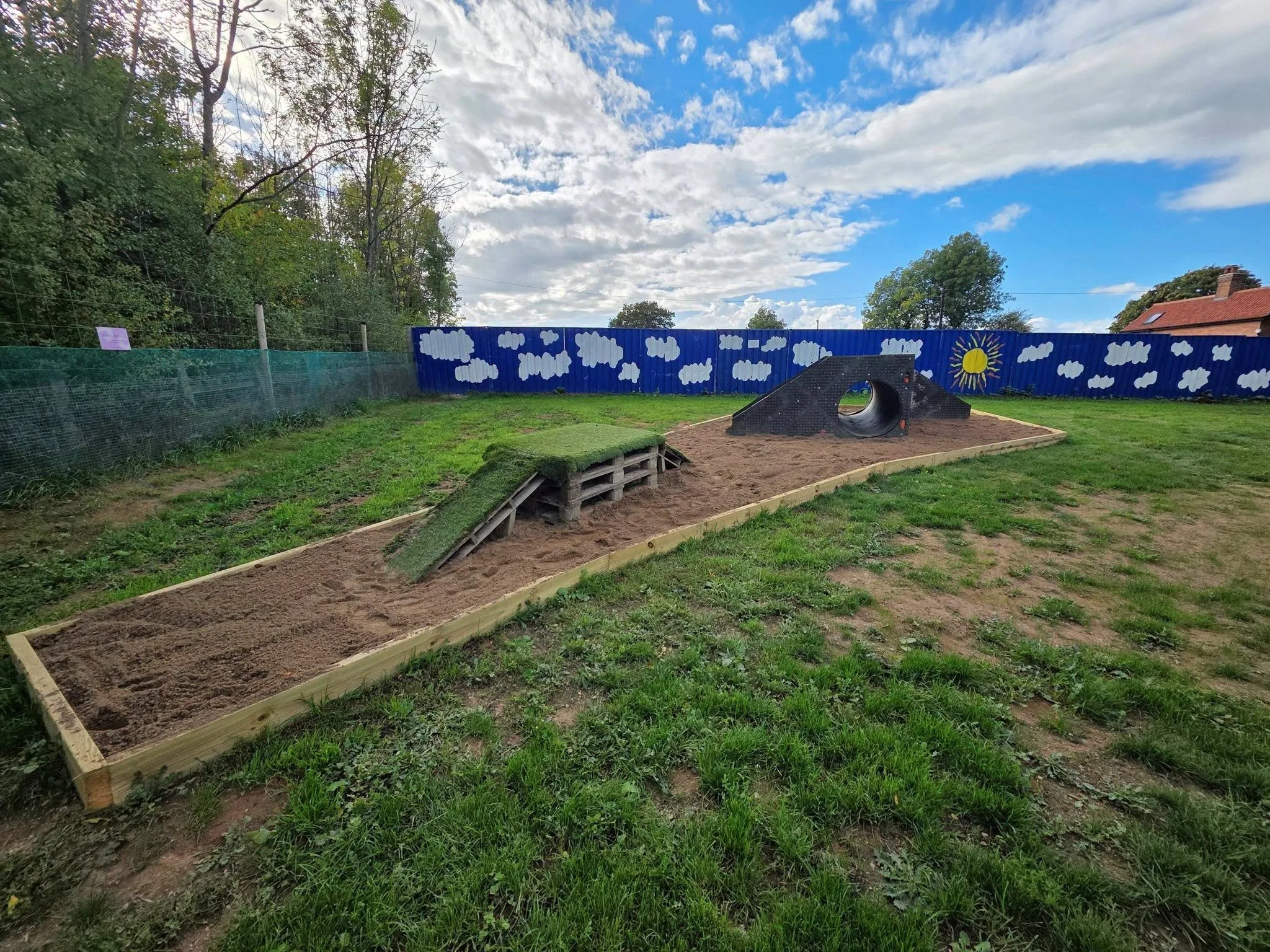 Small outdoor playground with painted blue fence depicting clouds, sun, and stars, surrounded by green trees at sunset.