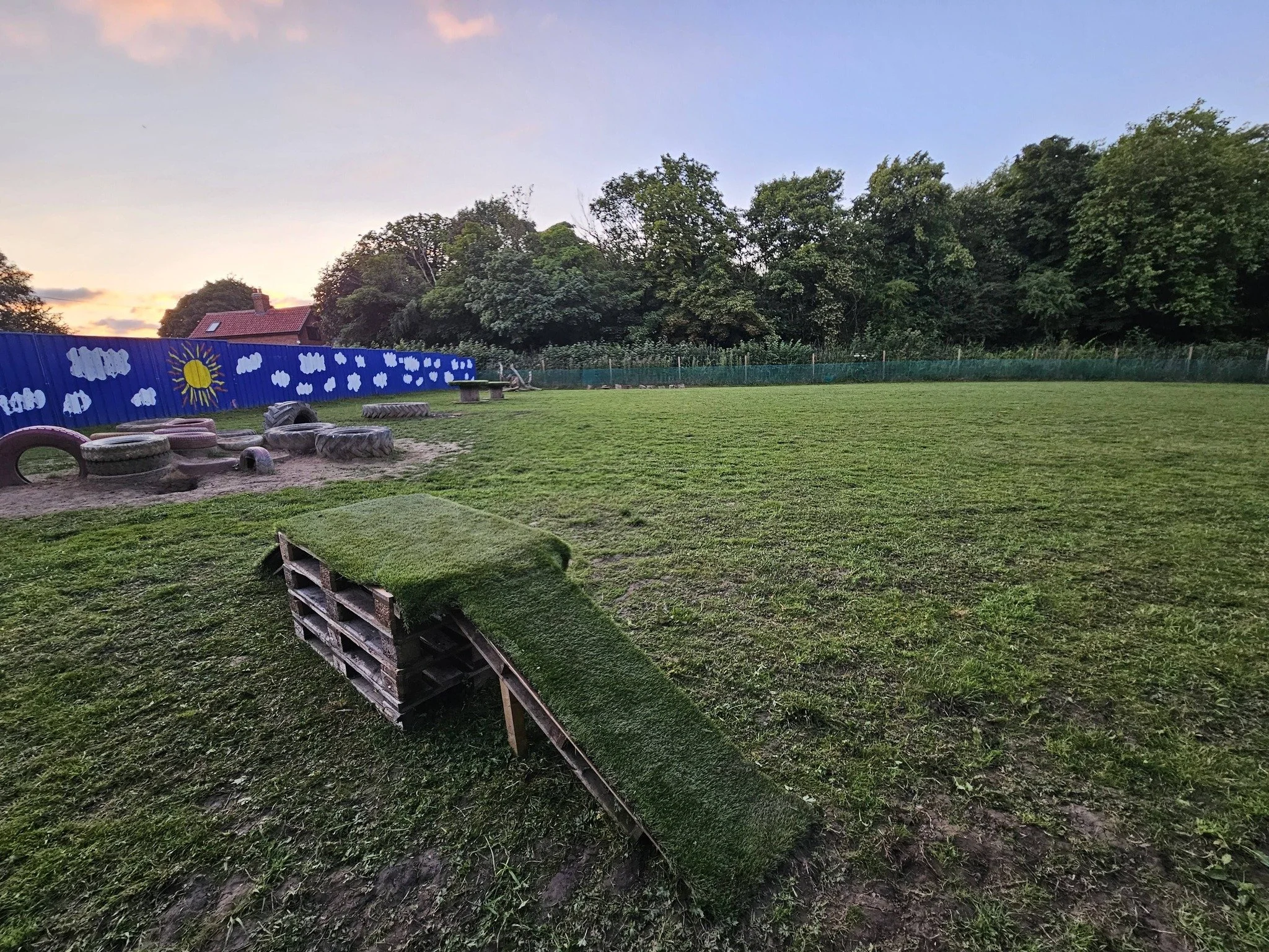 Small outdoor playground with painted blue fence depicting clouds, sun, and stars, surrounded by green trees at sunset.