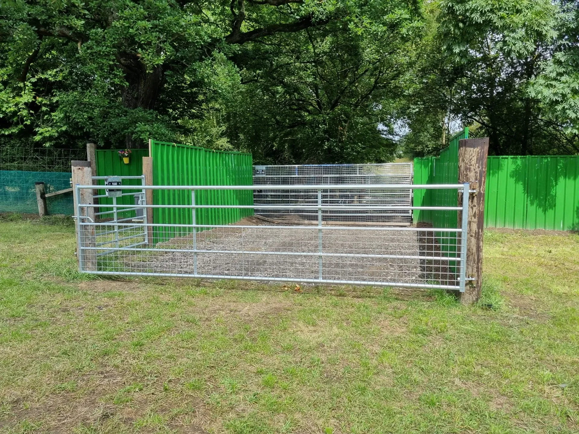 A green fenced enclosure with a metal gate, situated on a grassy area with trees in the background.