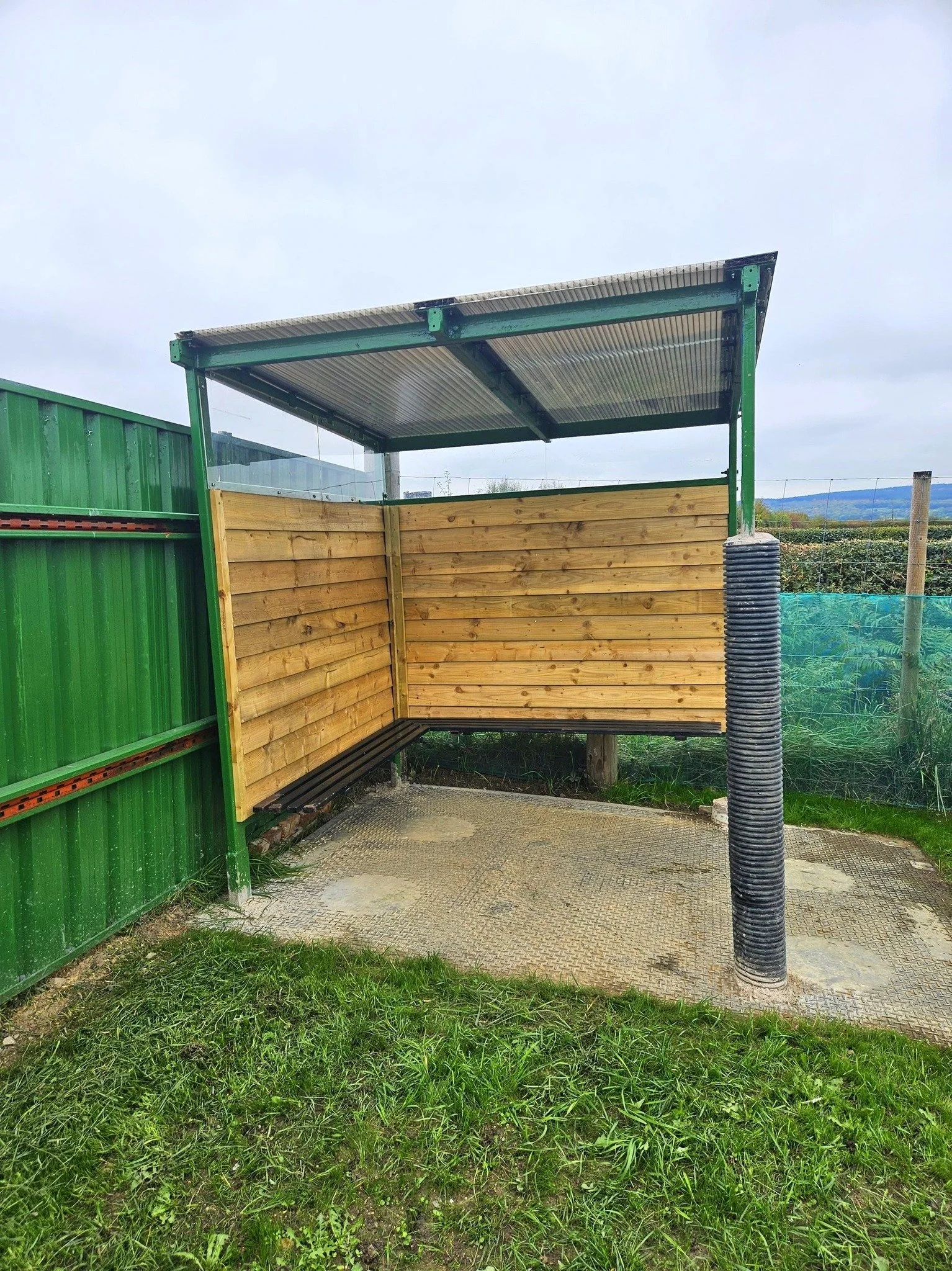 A partially constructed outdoor shelter with a metal roof, wooden side paneling, and black bench, situated on a paved area next to a green fence.