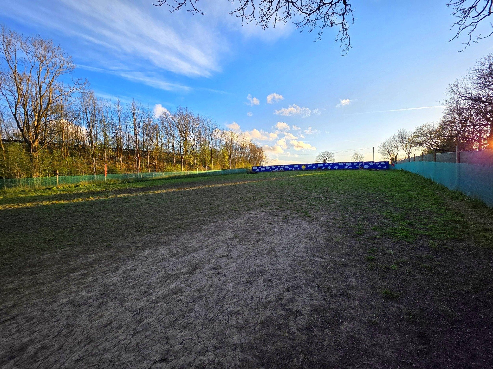 An empty dirt tennis court surrounded by a green fence, with bare trees and a blue sky with clouds in the background.