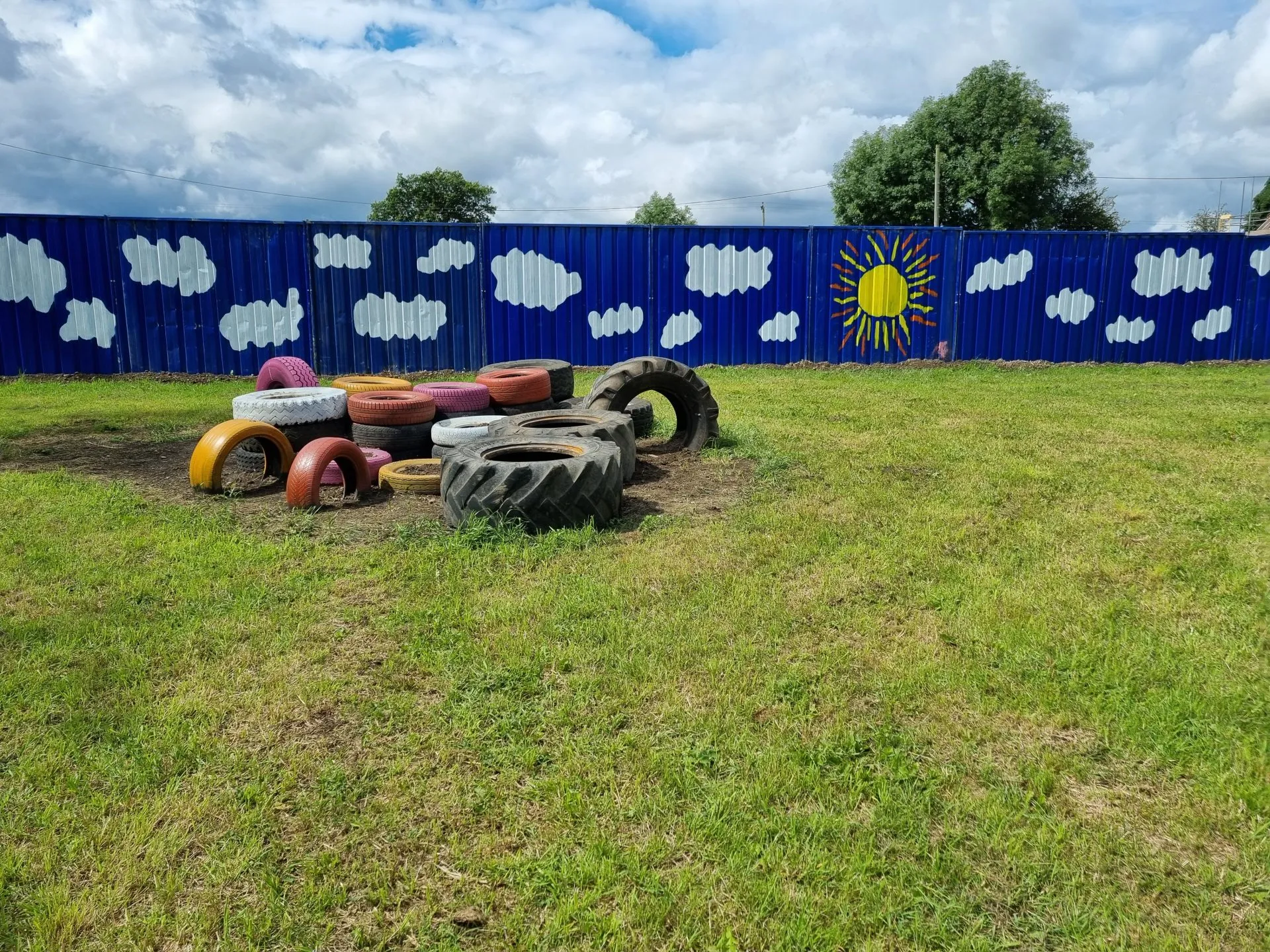 Colorful tires arranged in a small playground area in front of a blue fence decorated with clouds and a sun, with trees and cloudy sky in the background.