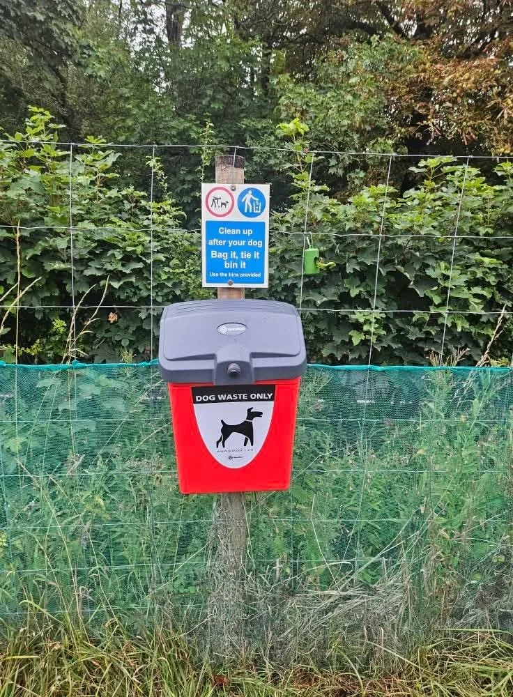A red dog waste disposal bin attached to a tree with a sign above it that instructs dog owners to clean up after their dogs, tie the waste bag, and use the provided bin. The background includes green foliage and a wire fence.