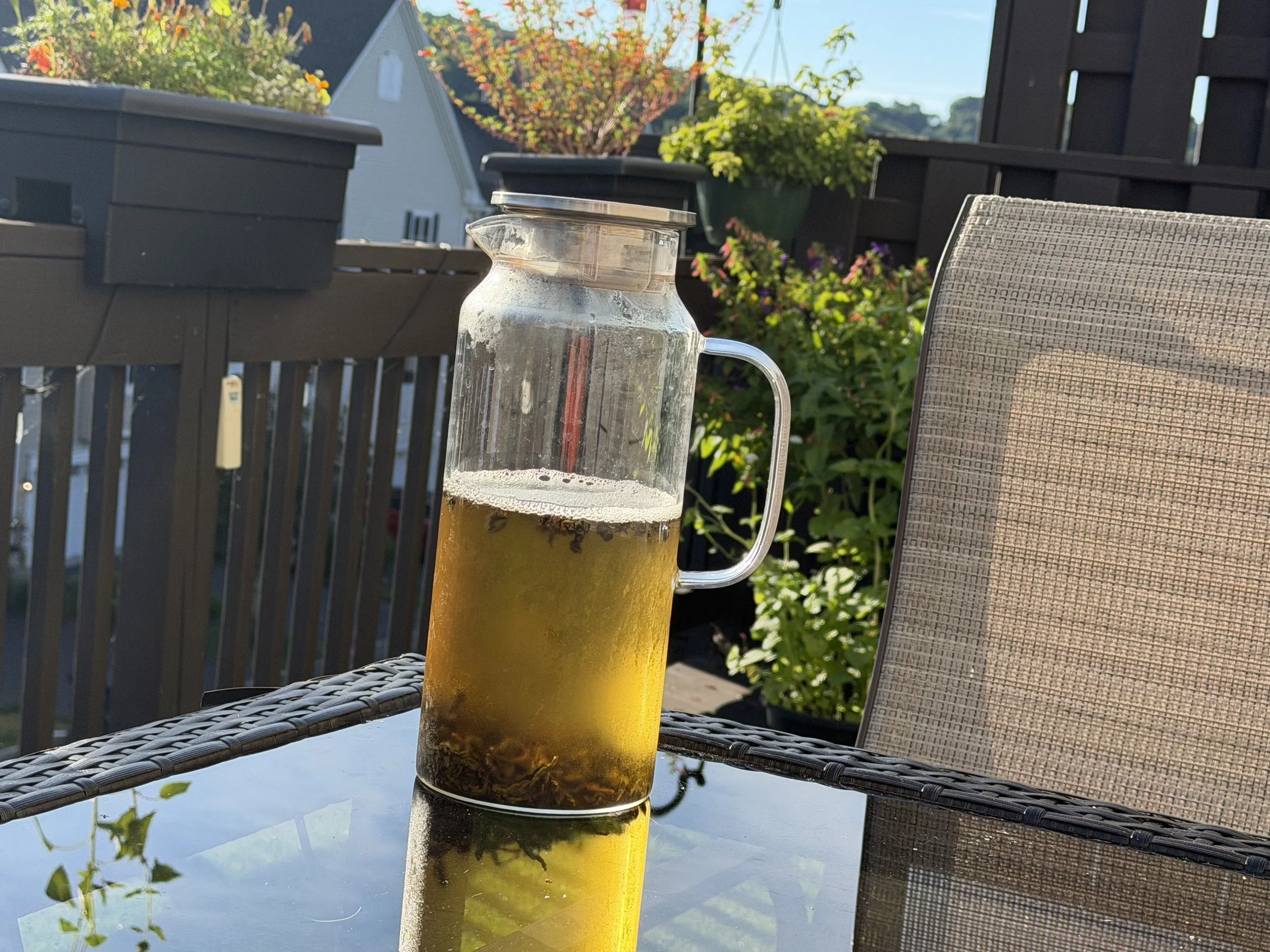 Glass pitcher of golden Iron Goddess of Mercy cold-brew oolong on a sunlit outdoor table surrounded by garden plants.