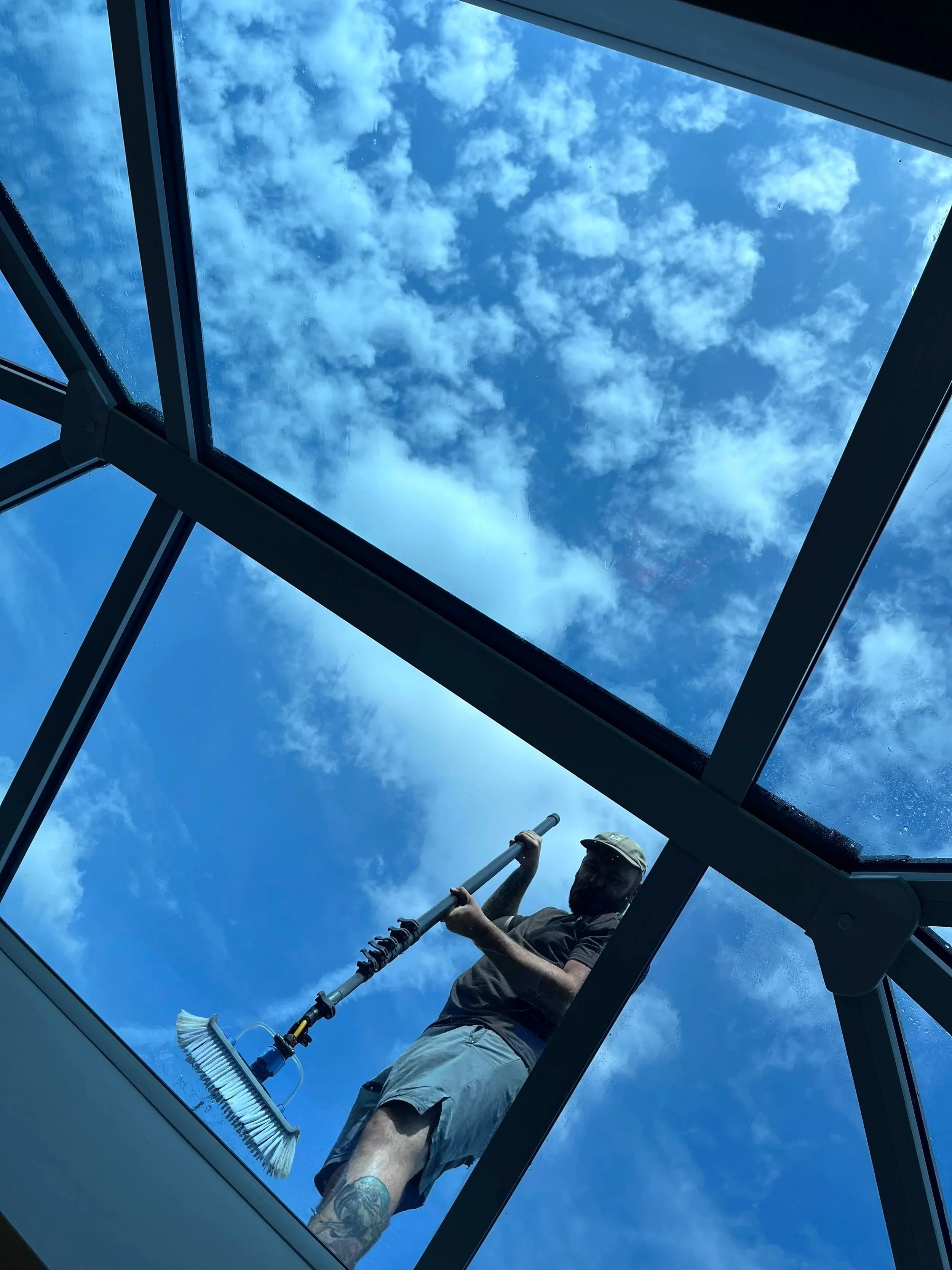 A person cleaning a glass ceiling or roof, seen from below with a blue sky and clouds above, holding a cleaning tool.