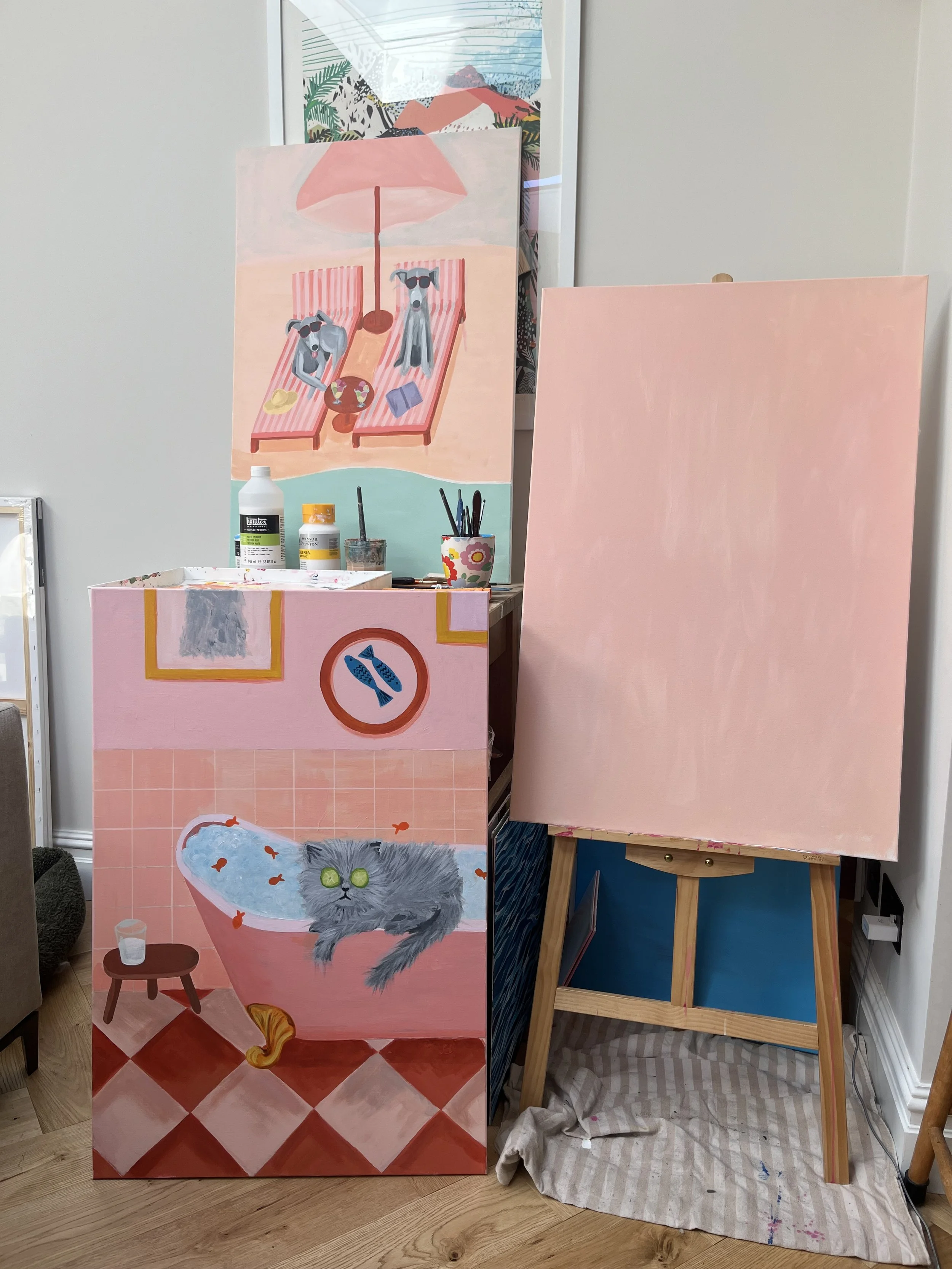 This image shows a partial view of a room with two pastel pink painted canvases, one on an easel and the other standing upright. The canvas on the left has a painting of a bathroom scene with a gray cat, a small table, and a fish bowl. The larger canvas behind it has a painting of a beach scene with two dogs lounging under a pink umbrella, with one wearing sunglasses. Art supplies and bottles are on a nearby table.