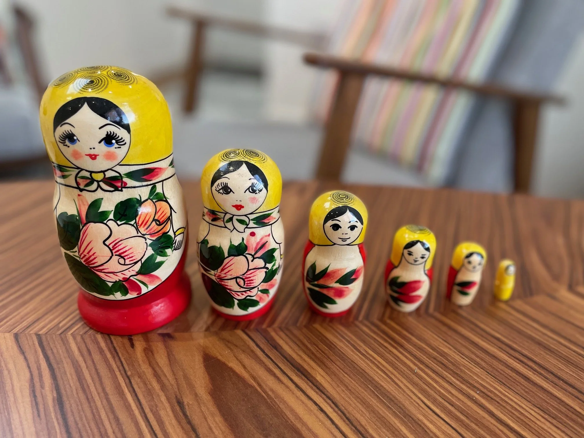 A set of six traditional Russian nesting dolls, or Matryoshka dolls, arranged from largest to smallest on a wooden surface. The dolls are painted with floral patterns and have yellow head coverings, with the largest doll featuring rosy cheeks and blue eyes.