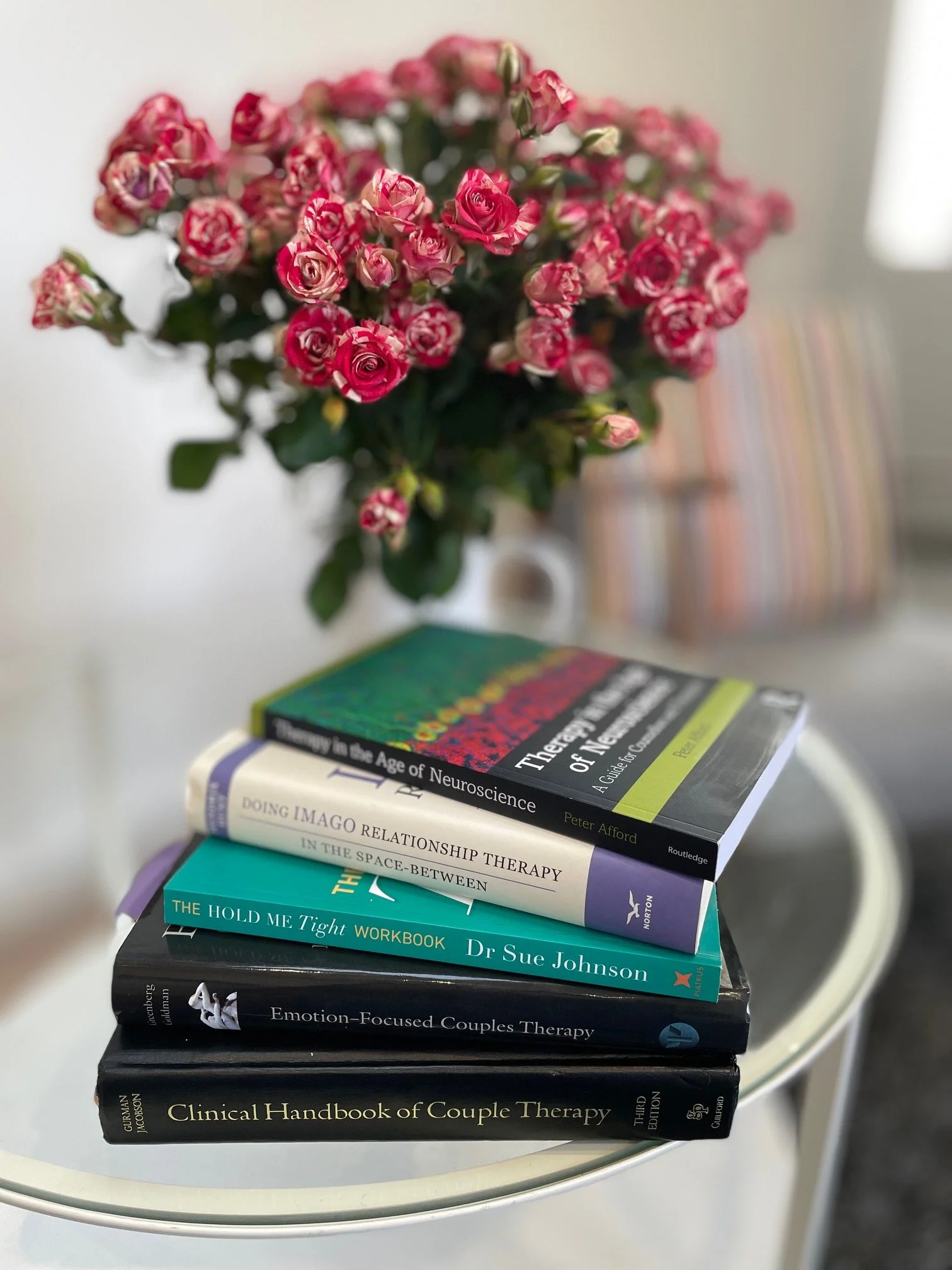 Stack of six books on a white table with a pink and white rose bouquet in the background and a striped chair visible in the distance.