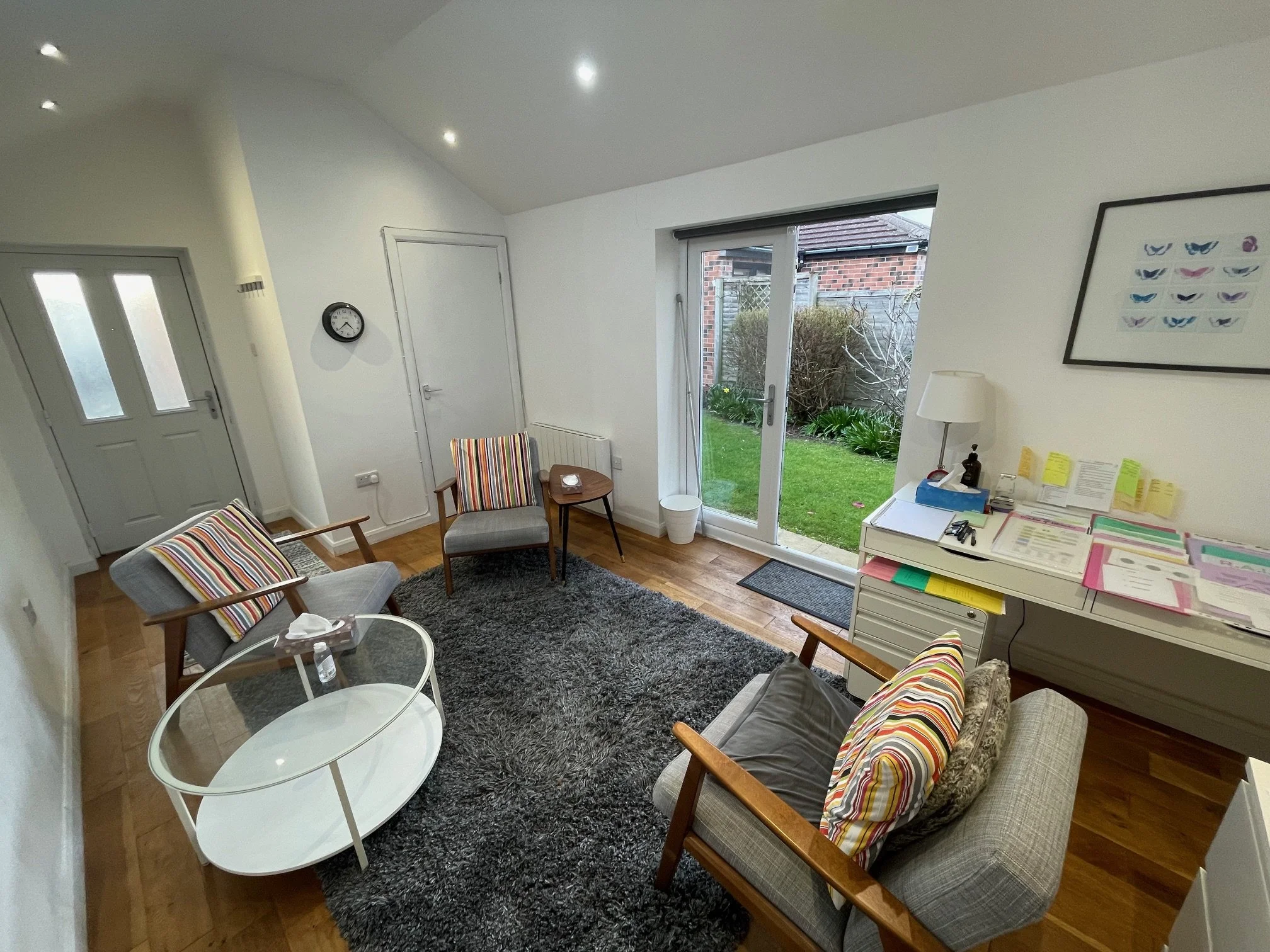 A cozy living room with a gray rug, wooden floor, and white walls. It has a gray door with windows, a small round table with a tissue box, two armchairs with striped cushions, a sliding glass door leading to a garden, a white desk cluttered with papers and office supplies, a lamp, and a framed butterfly artwork on the wall.