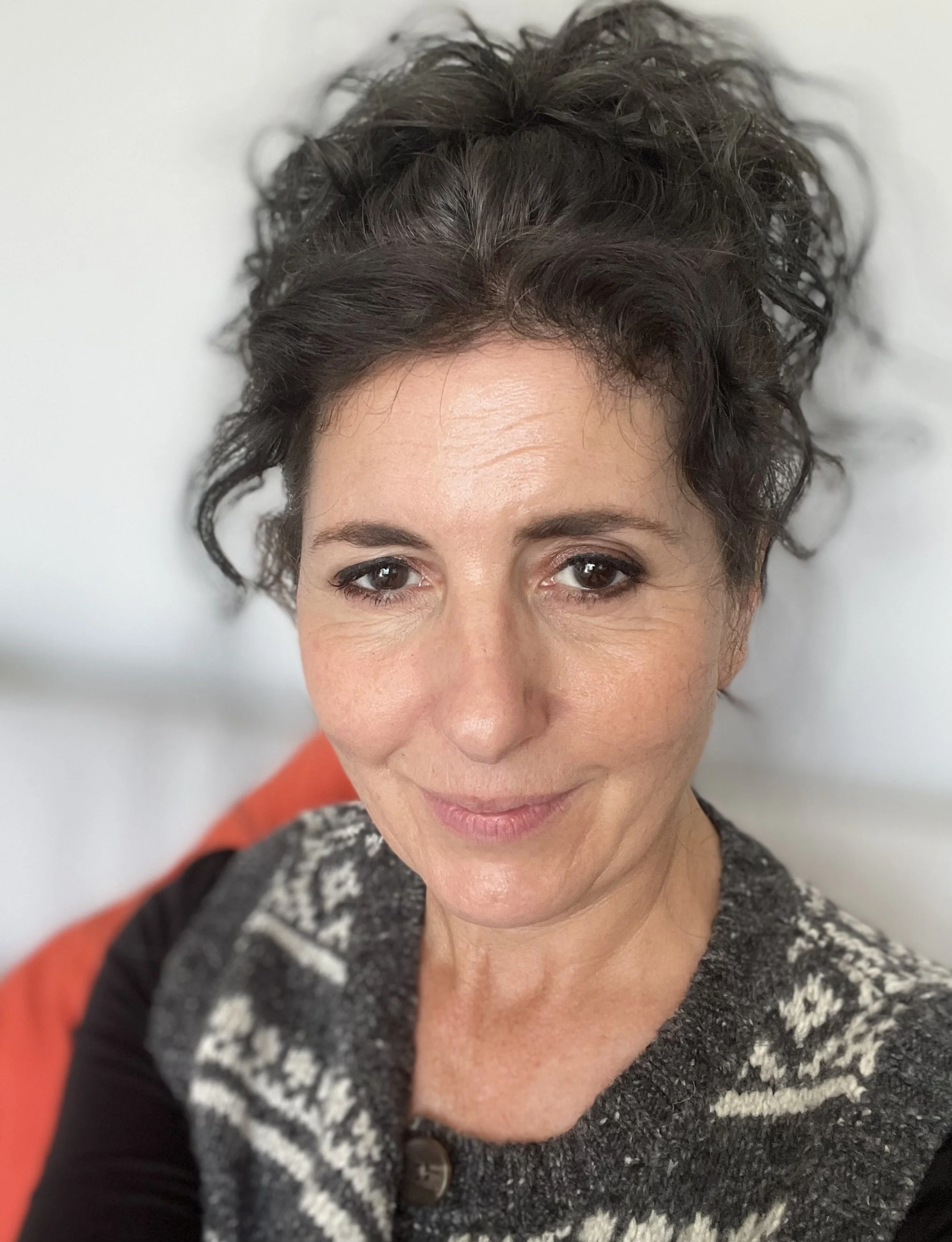 Close-up of a woman with short, curly brown hair and brown eyes, smiling slightly, wearing a patterned gray and black sweater and black top, seated against a light background.