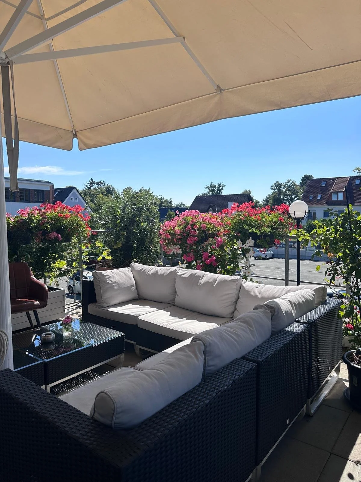 Geräumige Terrasse mit Lounge Sitzbereich und mehrfarbigen Blumen in Töpfen, unter Sonnenschirm, mit Blick auf Häuser und blauen Himmel.