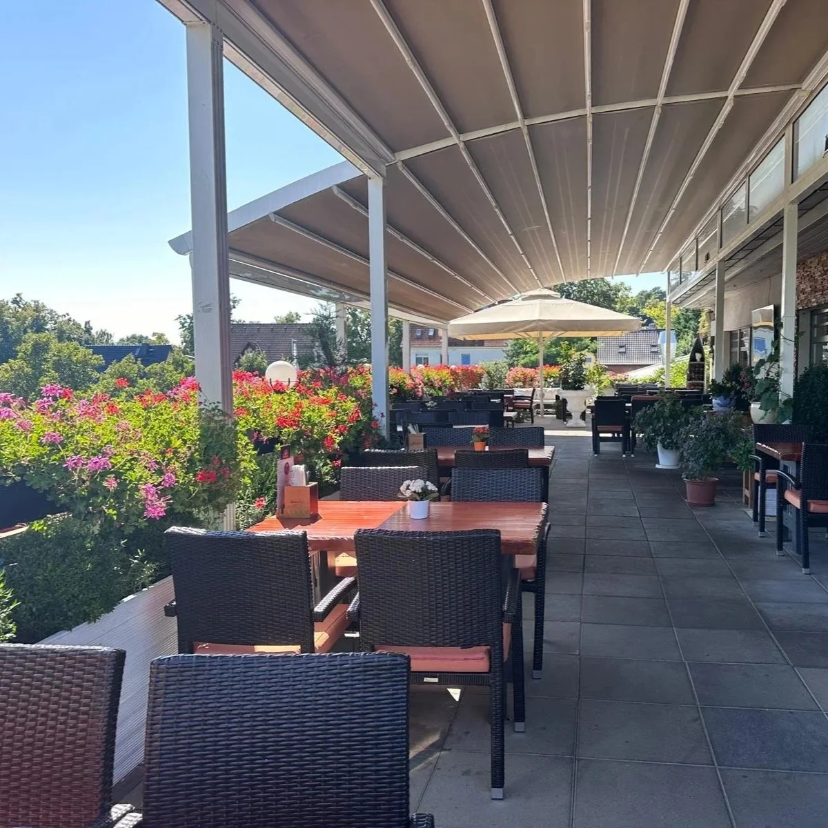 Außerhalb einer Terrasse mit Tischen und Stühlen, viele bunte Blumen und Pflanzen, Sonnenschirm, Himmel, freundliches Wetter.