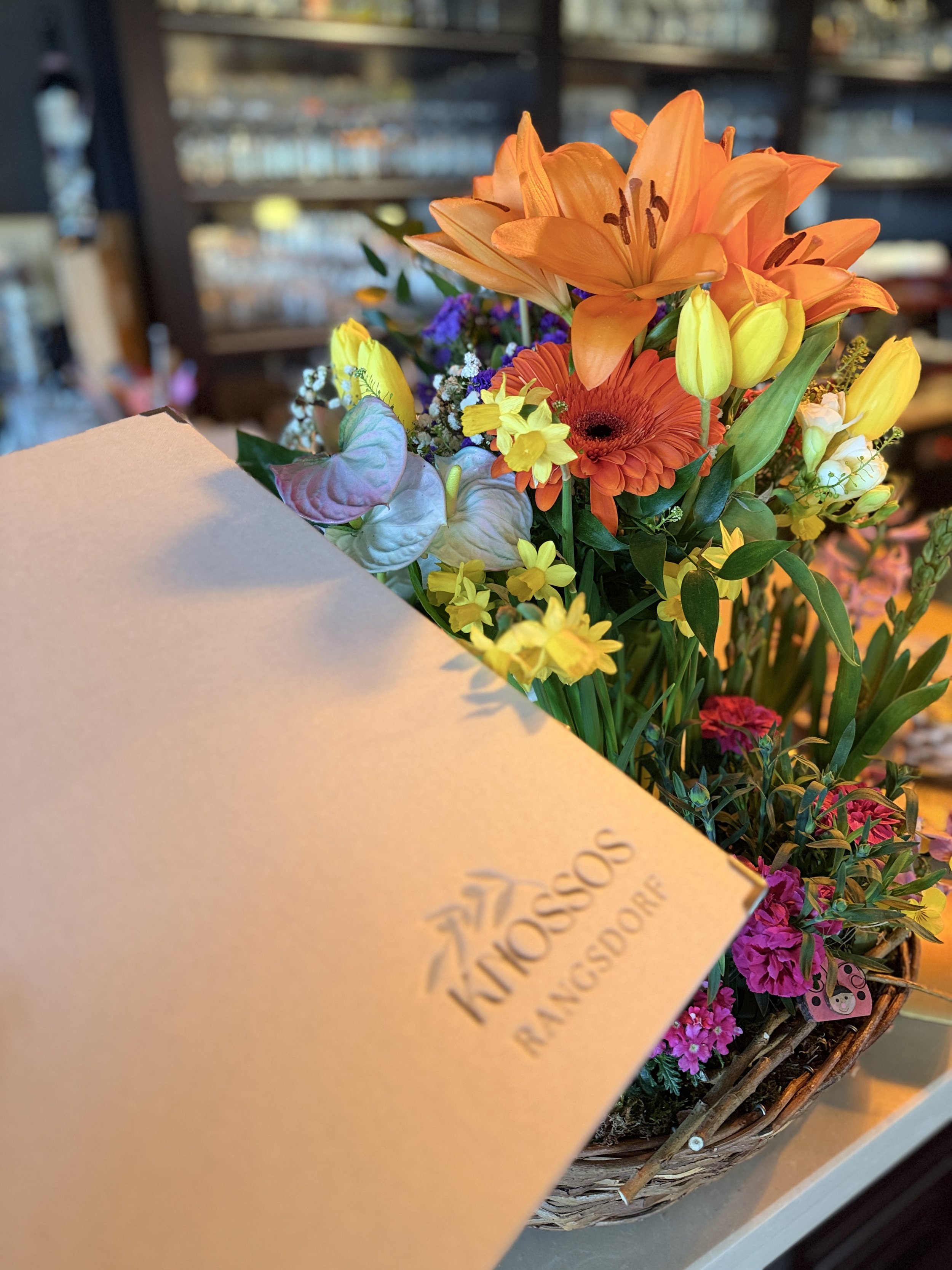 Bunte Frühlingsblumen, darunter orange Lilien, gelbe Trompetenblumen, rote Gerbera und kleine blaue Blumen, in einem Korb.