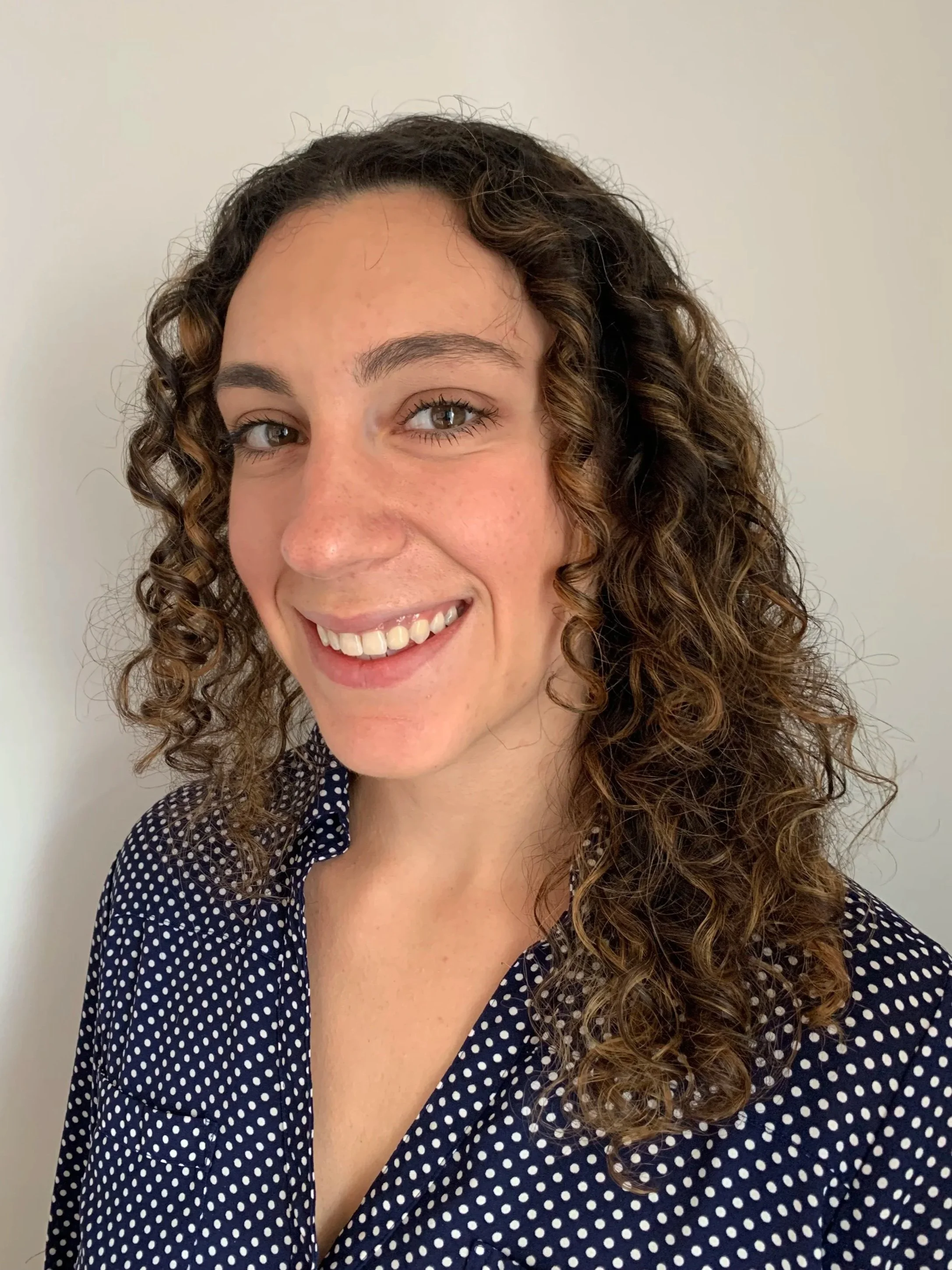 A woman with curly brown hair, light skin, and a bright smile wearing a polka dot shirt, standing against a plain light-colored wall.