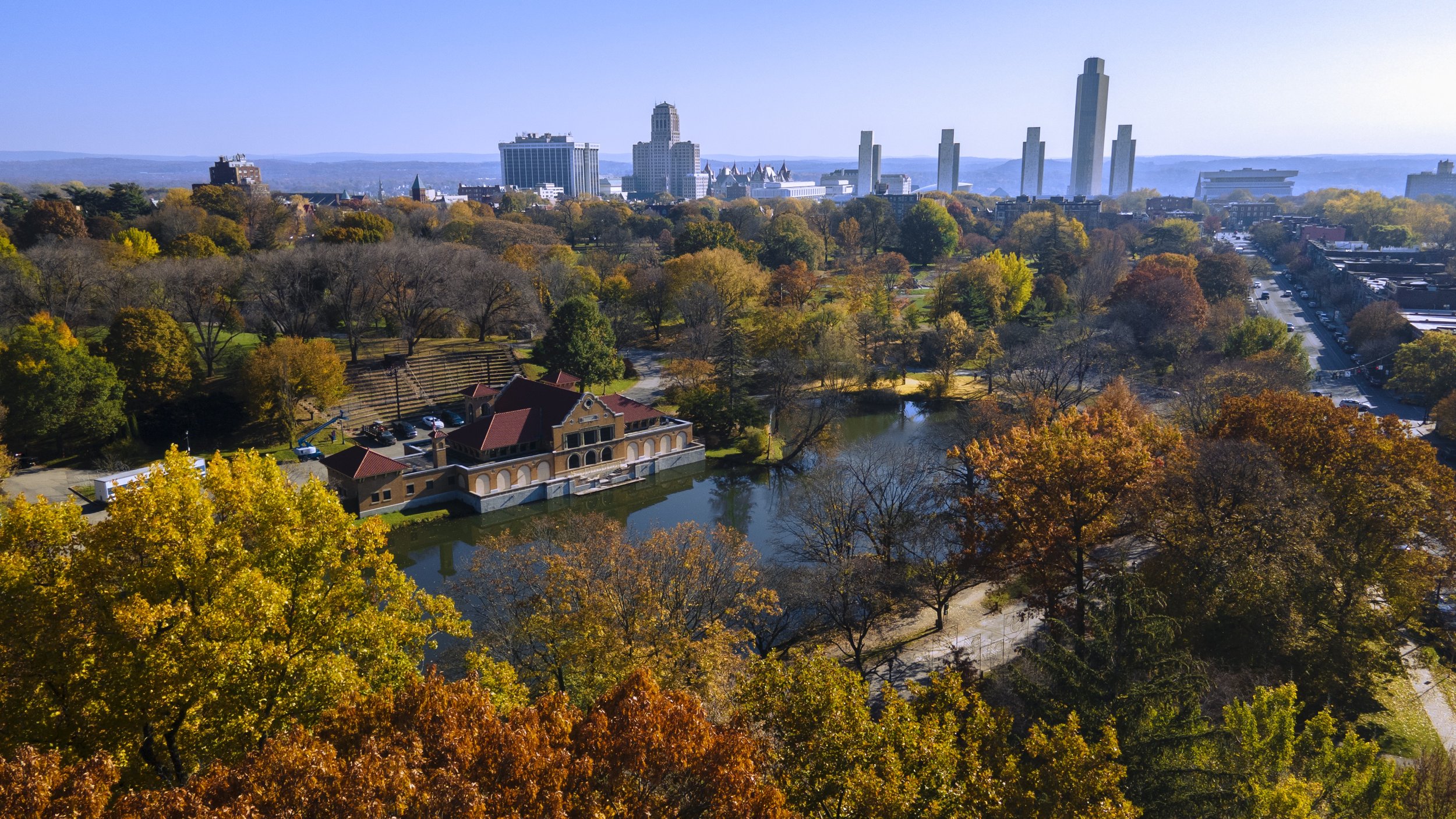 A city skyline with tall buildings in the distance and a park with colorful autumn trees and a small pond in the foreground.