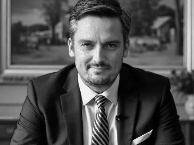 Black and white photo of a man in a suit and tie sitting at a desk, with a window showing trees in the background.