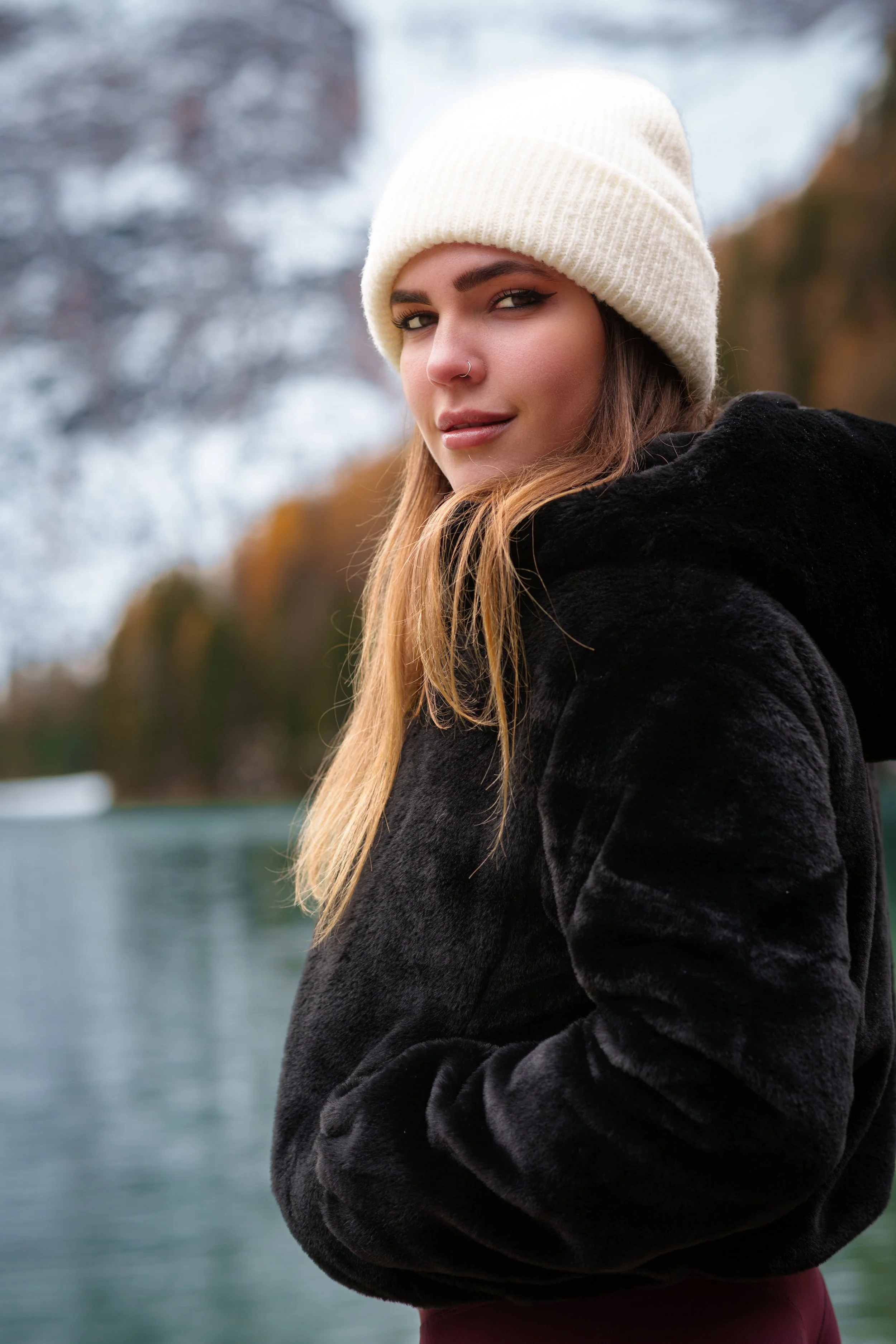Giada - Ragazza con cappello di lana bianco e cappotto nero al lago di Braies con acqua e alberi autunnali sullo sfondo.