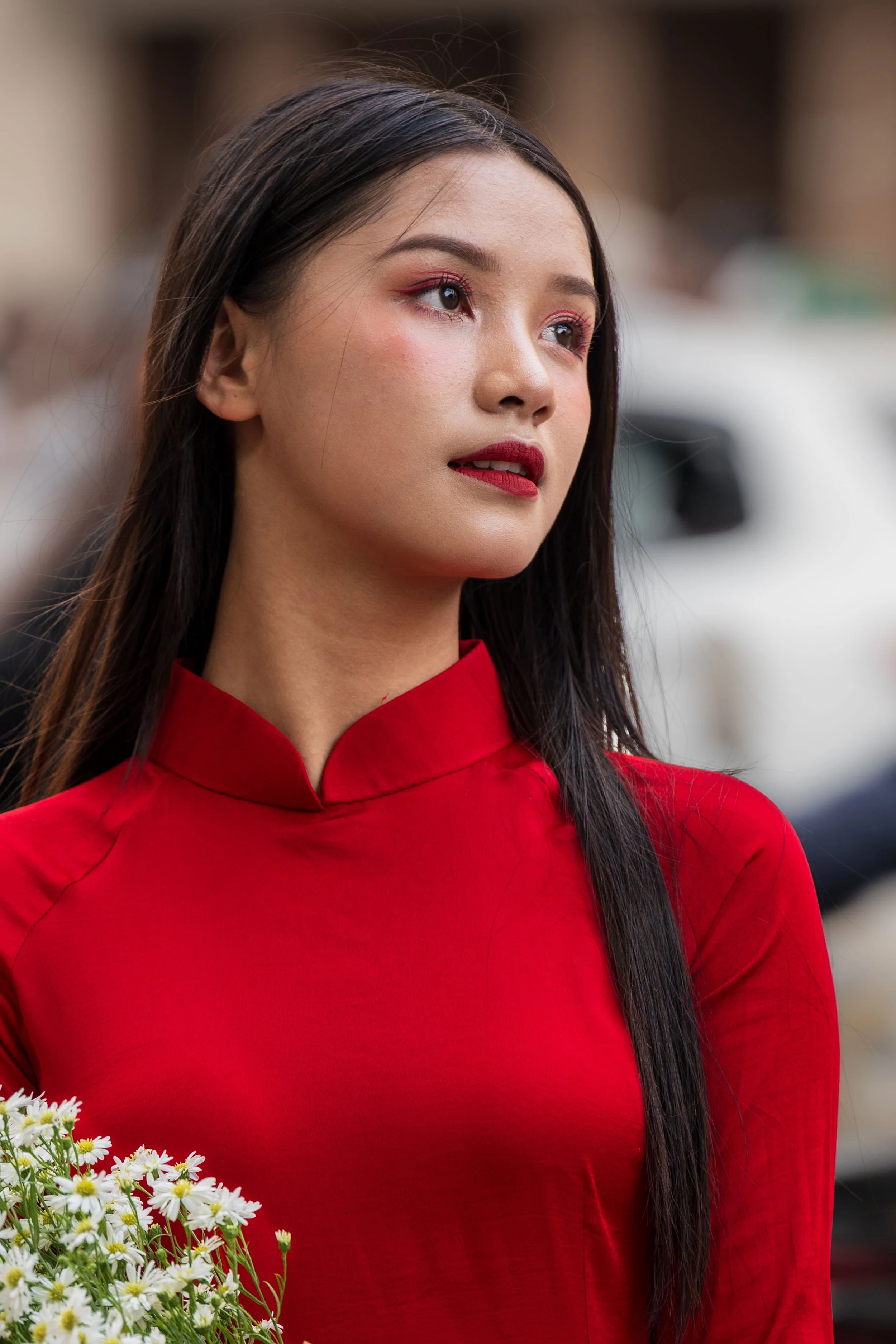 Vietnam - Ragazza con lunghi capelli castani, vestita con un qipao rosso, tiene un mazzo di margherite, con sfondo sfocato.