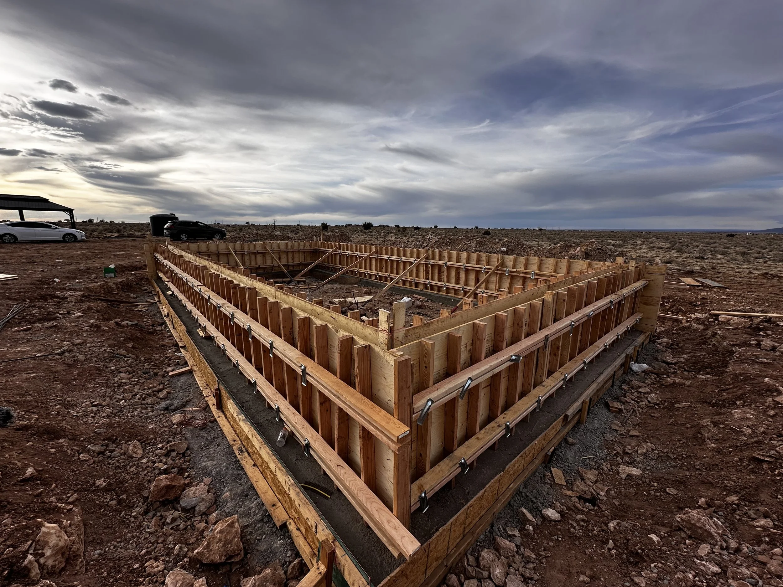 Construction site with wooden formwork for a building foundation in a desert landscape, under cloudy sky.
