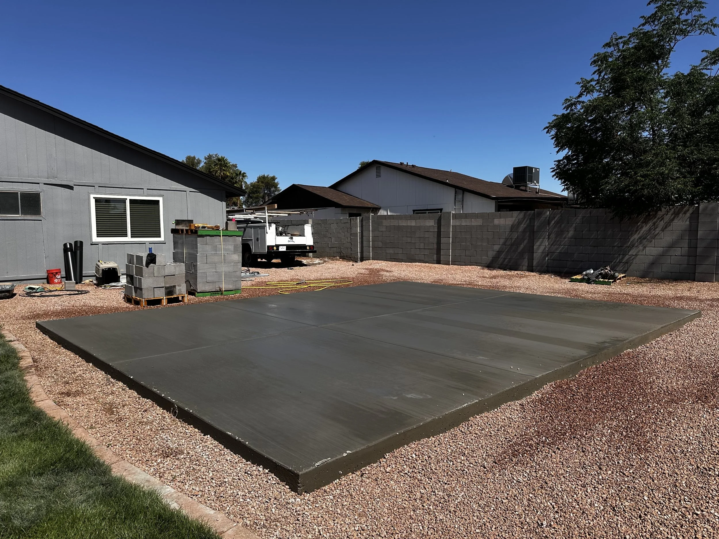 Backyard with a newly poured concrete slab, surrounded by gravel, with construction tools and equipment, a gray house, and a tall cinder block fence under a clear blue sky.