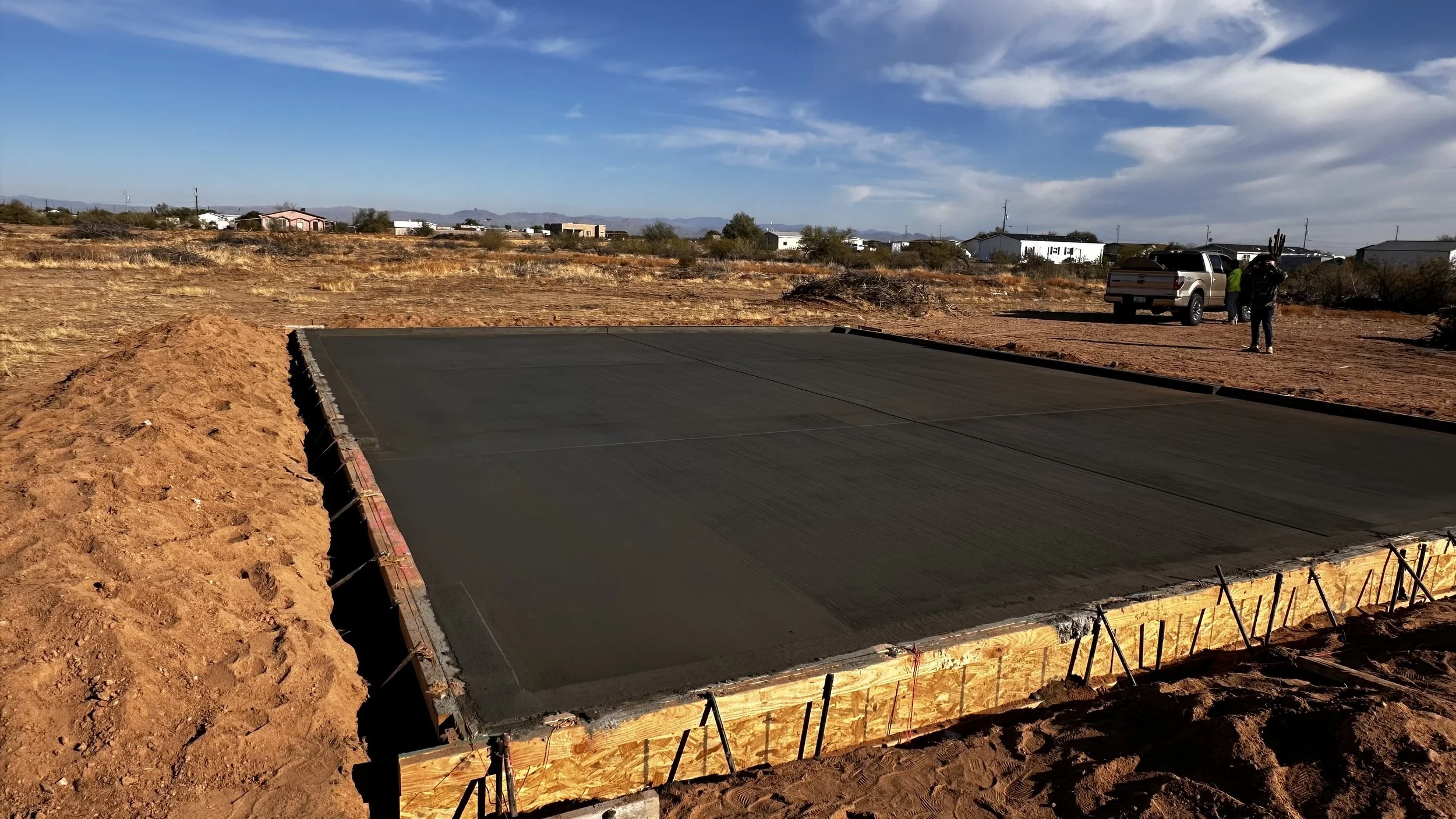 Concrete foundation being poured at a construction site with a dirt field, vehicles, and buildings in the background under a partly cloudy sky.