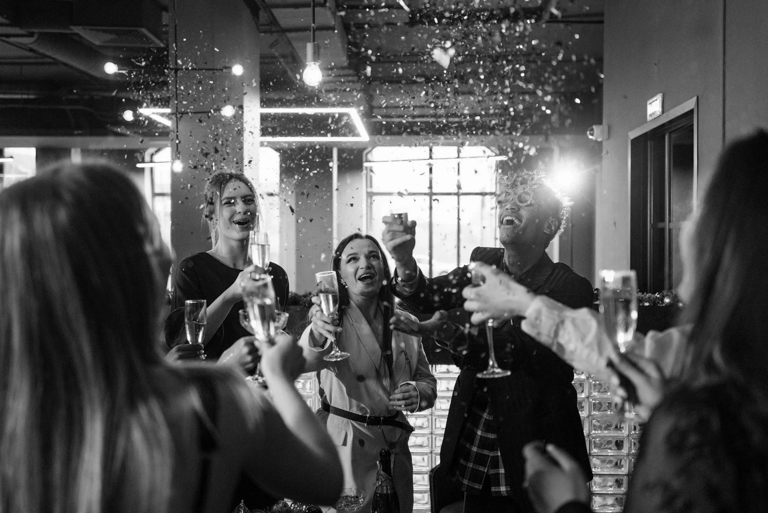 Group of people celebrating with drinks at a party, some wearing glasses, with confetti falling, in a modern indoor setting.