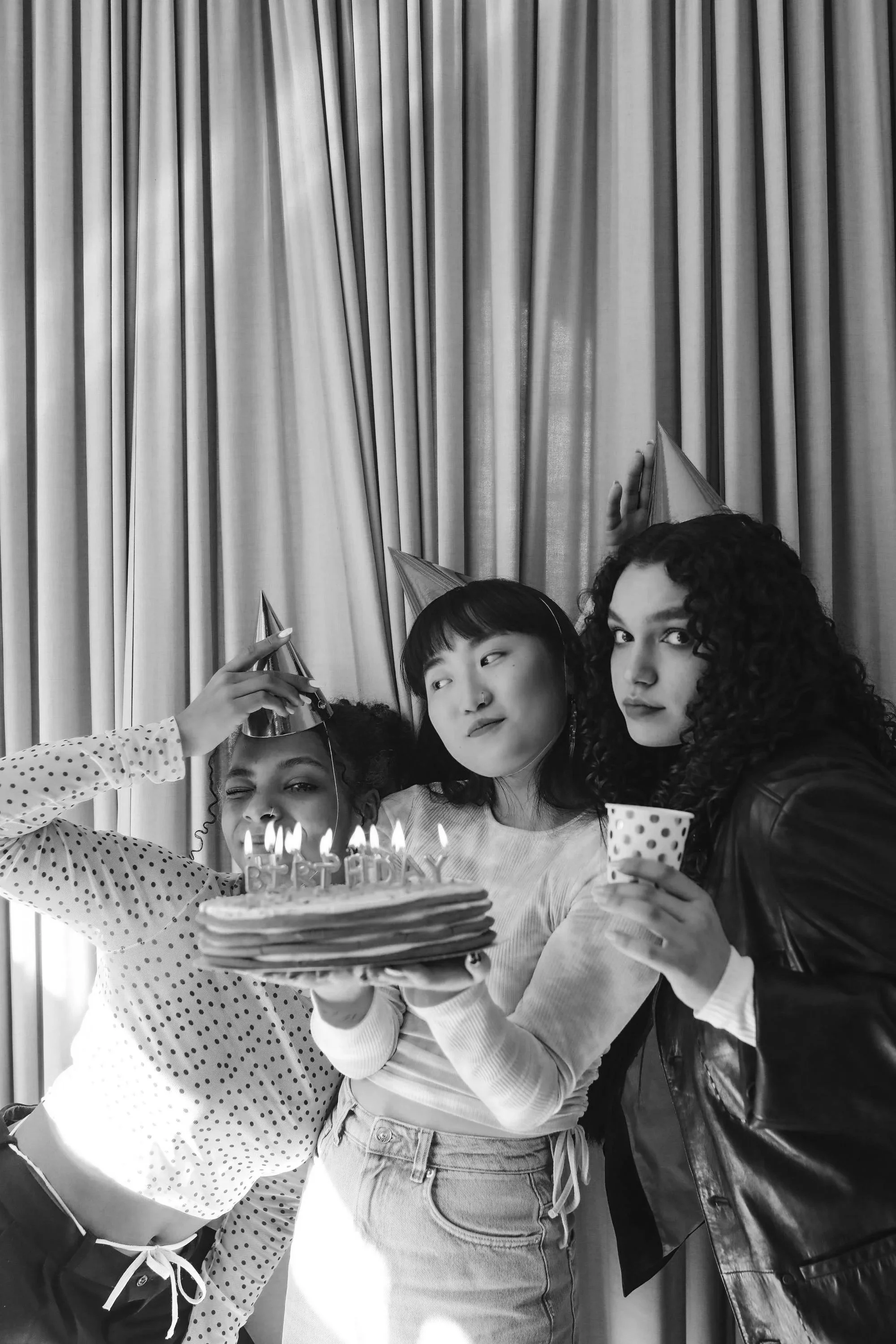 Three young women celebrating a birthday in front of a curtain, with a birthday cake with lit candles, party hats, and a polka dot cup.