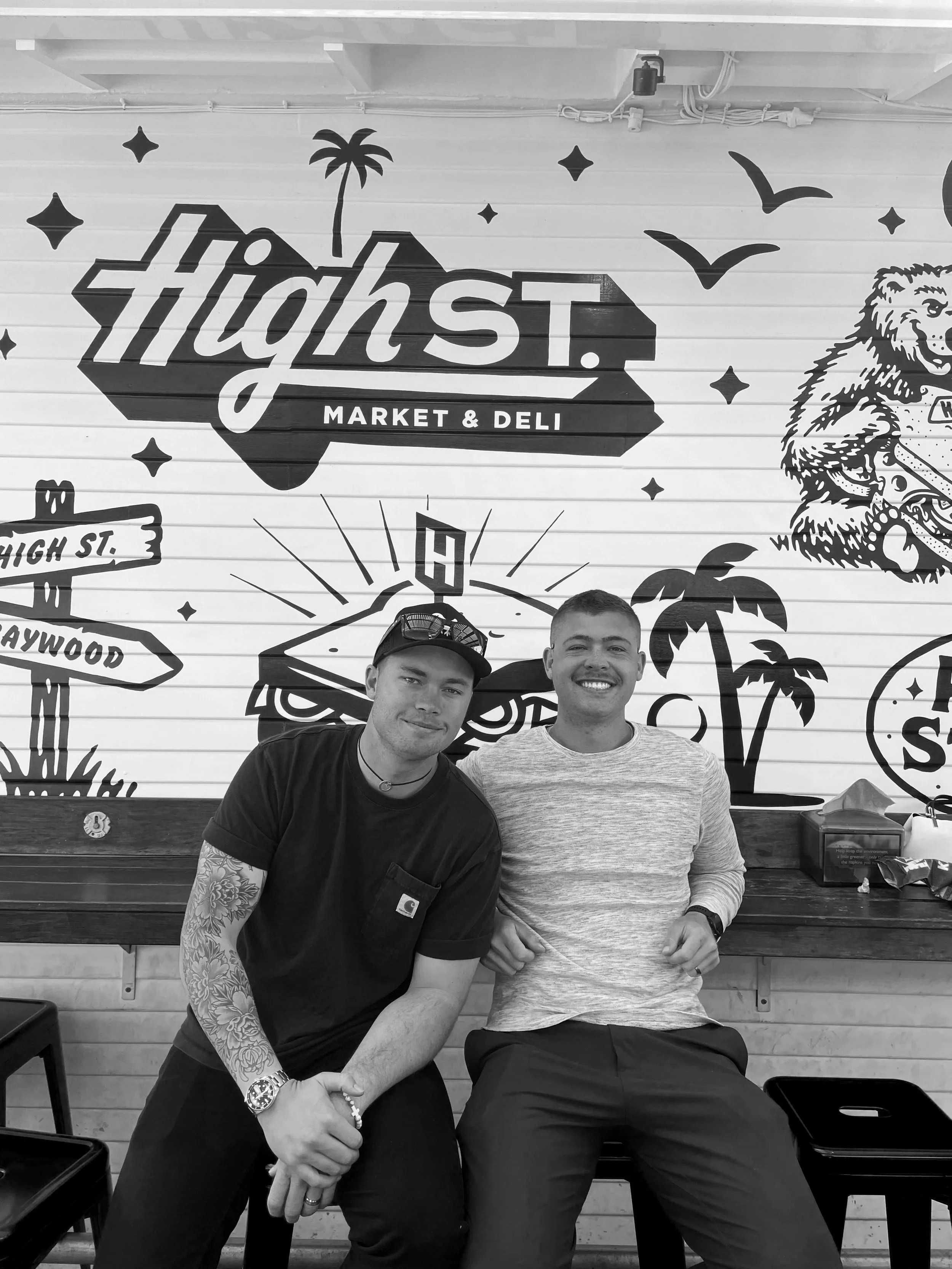 Two young men sitting and smiling in front of a wall with a mural that reads 'High St. Market & Deli,' featuring palm trees, birds, and other decorative illustrations.