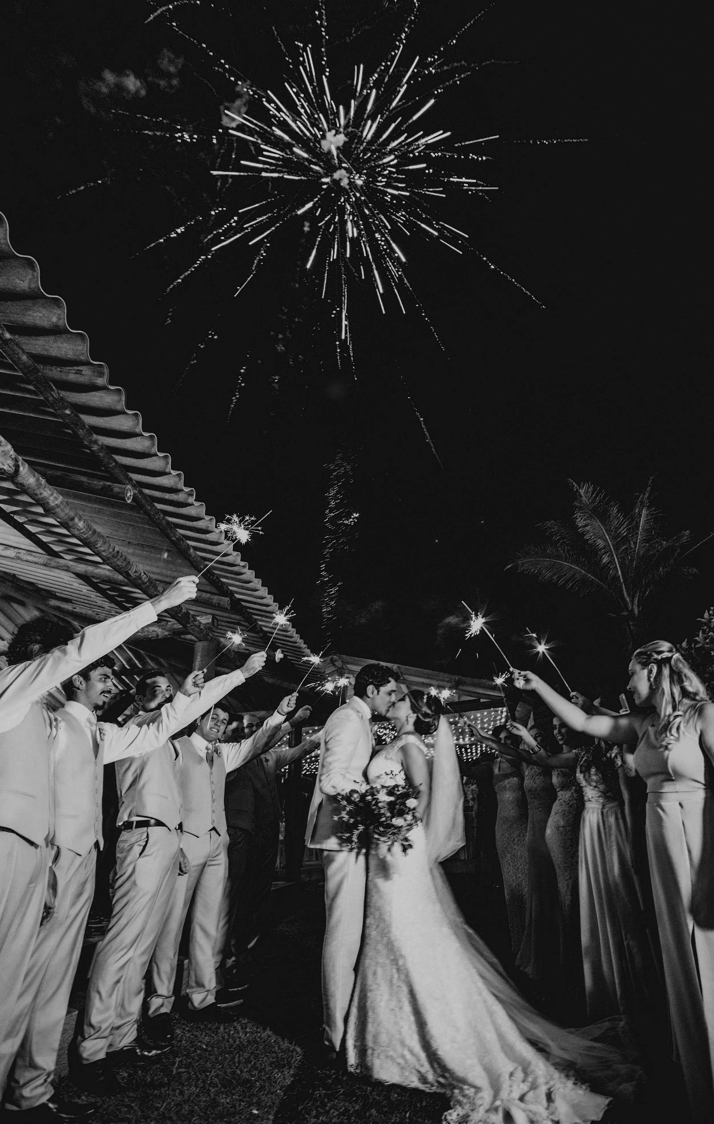 A black and white photo of a wedding celebration at night, with a couple kissing in the center, surrounded by friends holding sparklers. Fireworks light up the sky above.