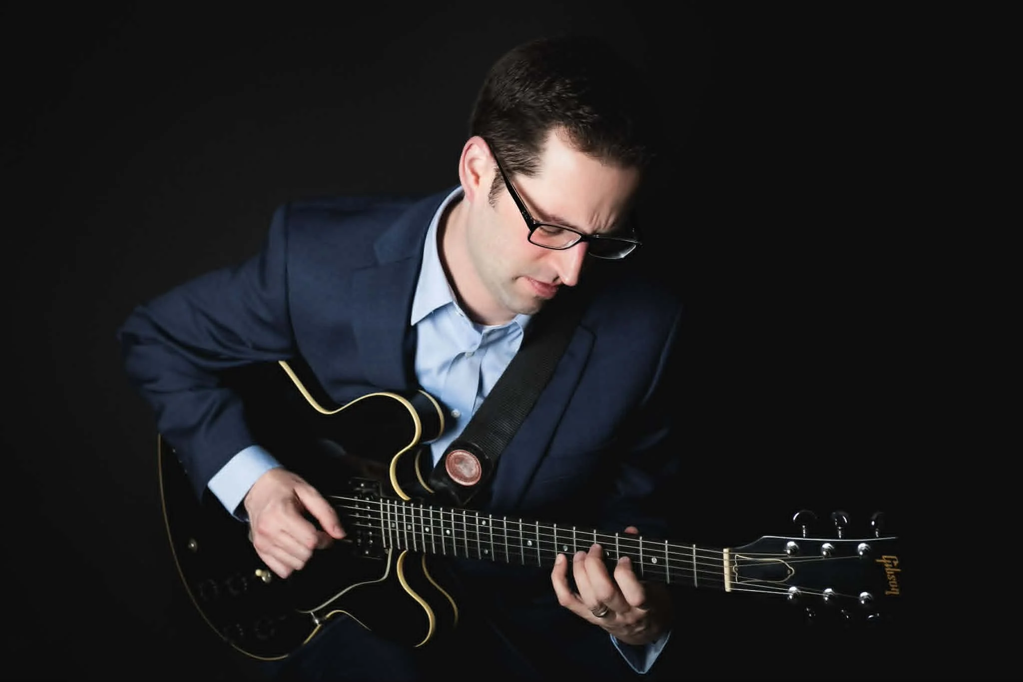 A man in a suit and glasses playing an acoustic guitar against a black background.