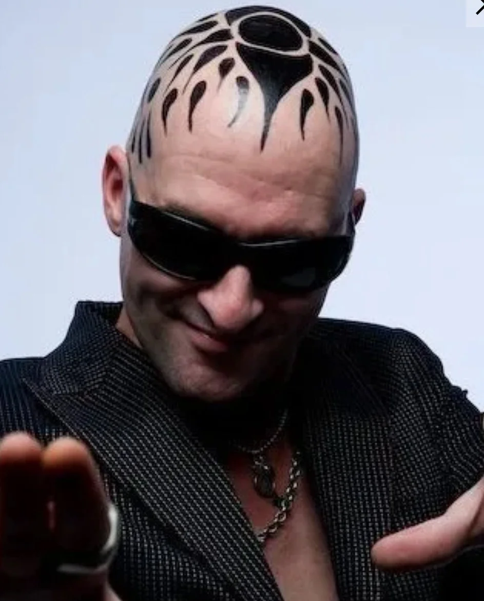 A man with a shaved head featuring black tribal-style hair tattoos, wearing sunglasses, a black textured blazer, and layered necklaces pointing towards the camera.