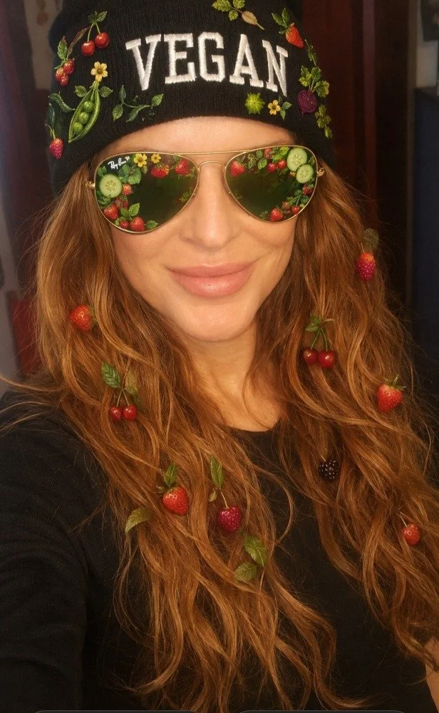 A woman with long red curly hair wearing a black beanie with colorful embroidery and the word 'VEGAN' on it, gold sunglasses reflecting strawberries, cherries, and cucumbers, and strawberry and cherry accessories in her hair.