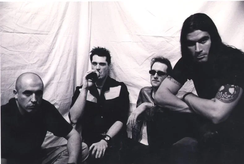 Black and white photo of four men sitting together against a plain backdrop, some with sunglasses and tattoos, with a serious expressions.