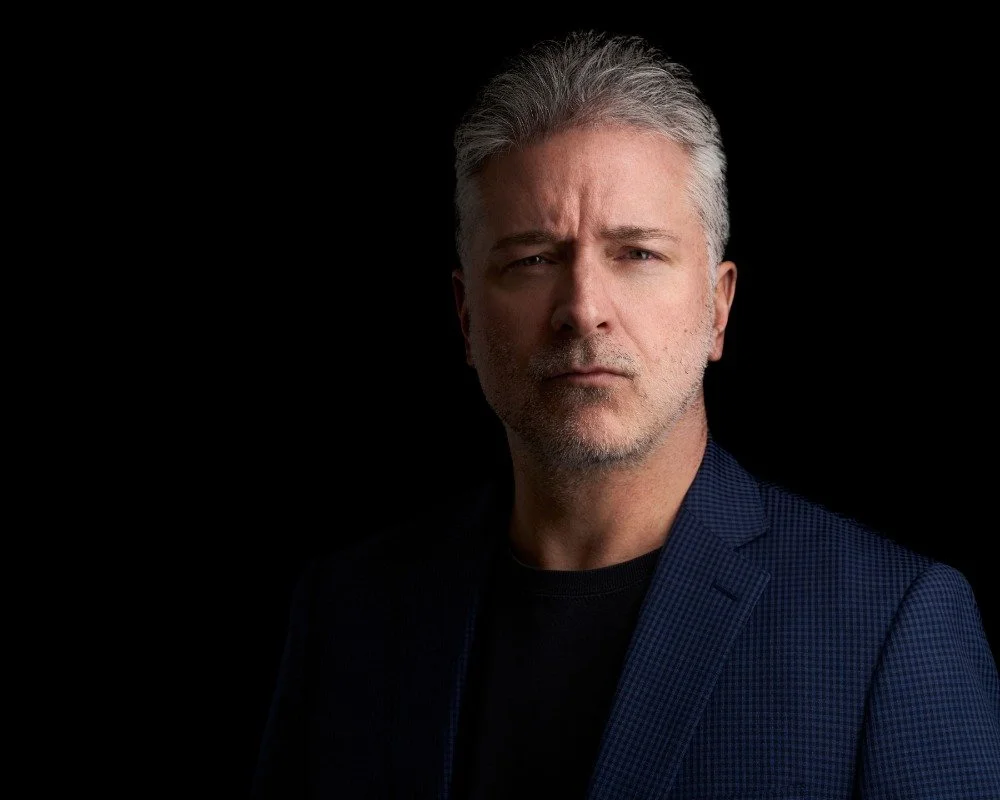 Portrait of a middle-aged man with gray hair and beard, wearing a dark blazer and black shirt, against a black background.