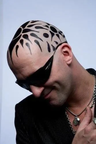 Person with shaved head and black tribal-style hair design, wearing sunglasses and jewelry, smiling with head tilted down.