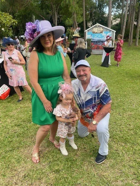 A woman in a green dress and large purple hat with flowers stands next to a man in a colorful short-sleeved shirt and cap, both smiling, with a young girl holding a small pet. Other people are in the background at an outdoor event on a grassy area wi