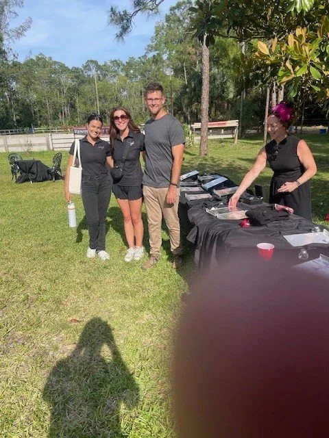 Three people standing outdoors on a grassy area near a table with food and drinks, with another person behind the table. There is a black dog in the background and trees surrounding the scene.