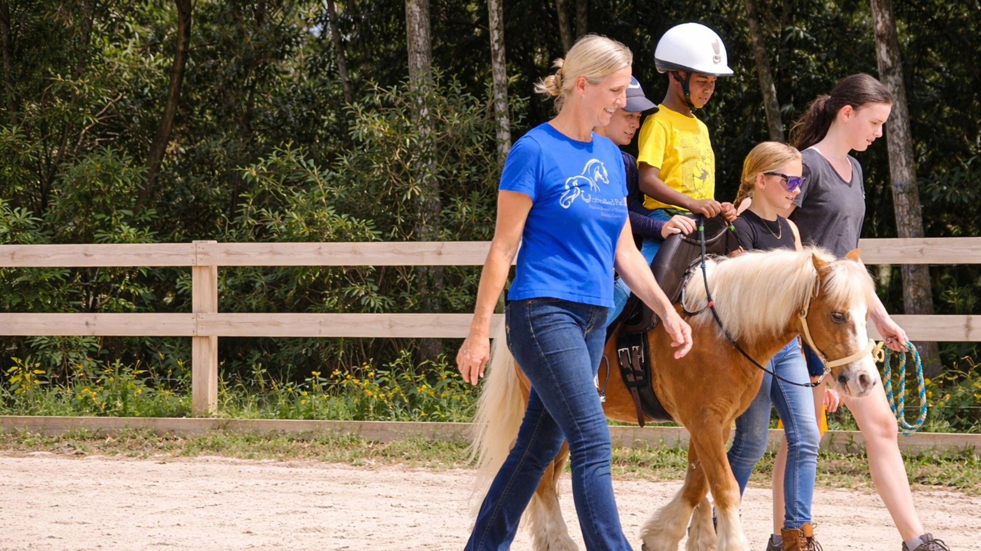 A woman leading a young girl who is riding a small horse in an outdoor riding arena with a wooden fence and trees in the background.