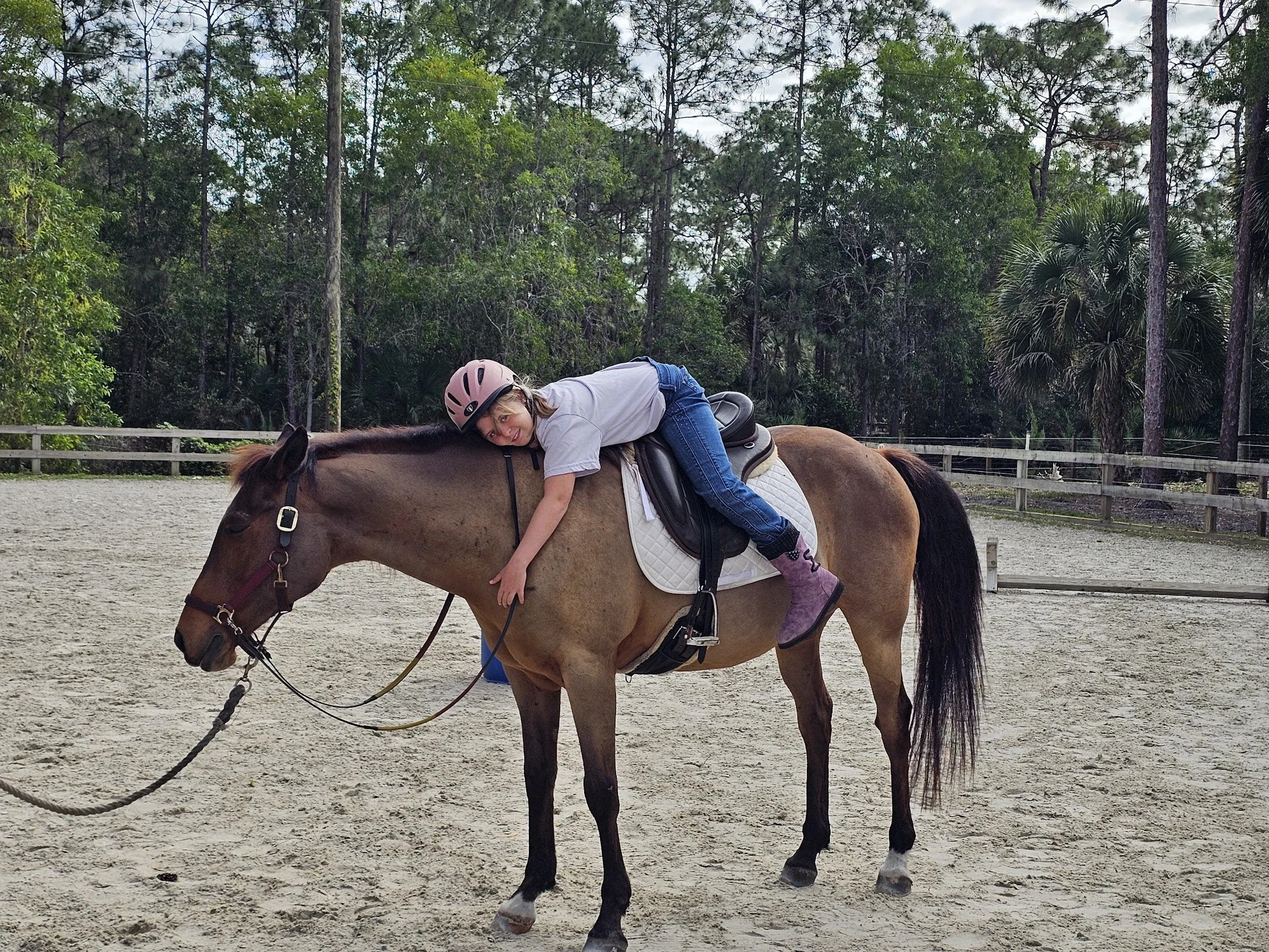 A girl wearing a pink helmet, white T-shirt, blue jeans, and pink boots is lying on the back of a brown horse with a black mane and tail, in an outdoor riding arena with trees in the background.