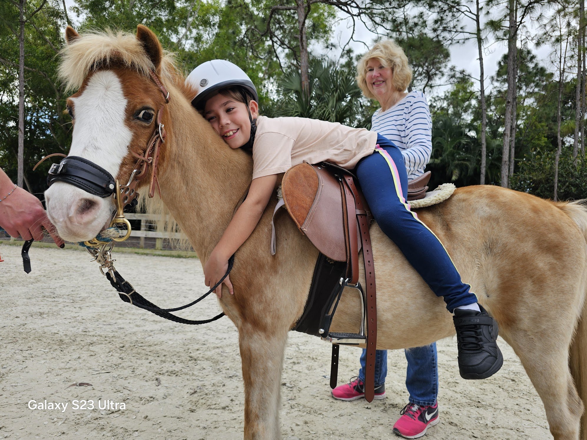 A young boy and an older woman riding a light brown horse with a white face at an outdoor equestrian facility, surrounded by trees.