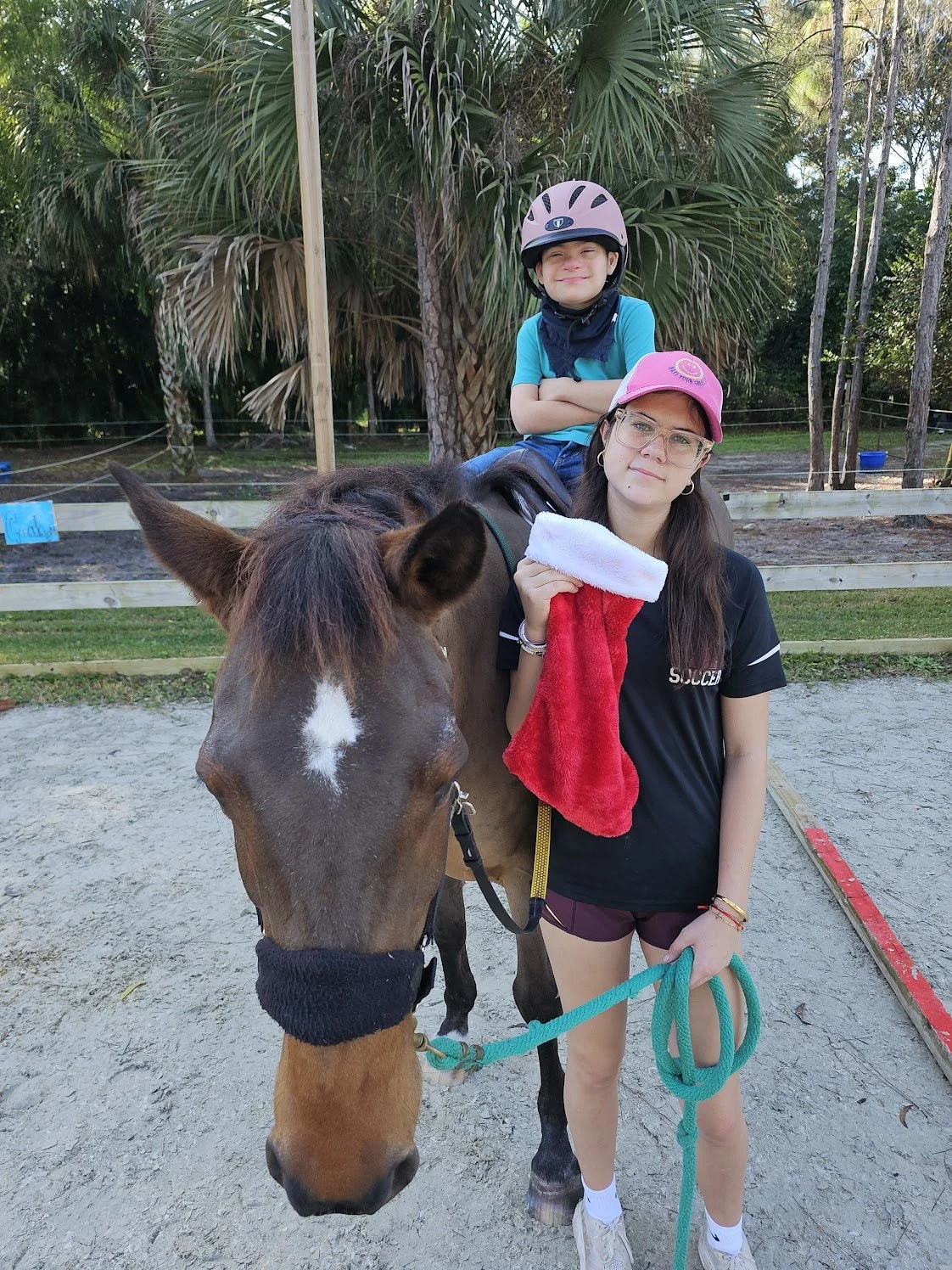 A young girl standing beside a brown horse holding a red and white Christmas stocking, with a boy sitting on the horse's back, in an outdoor setting with palm trees in the background.