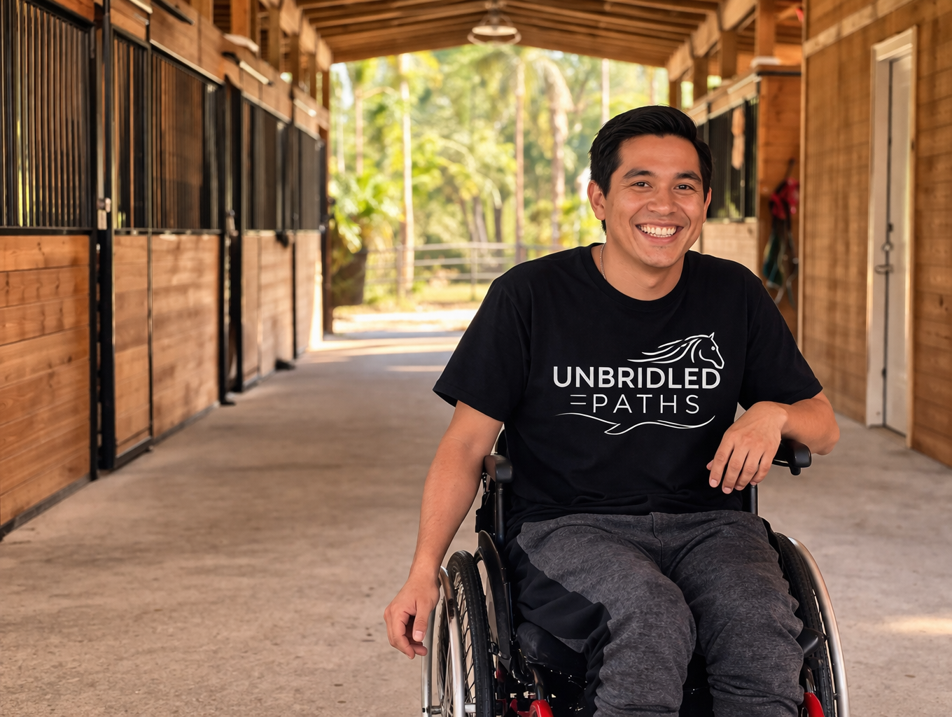 A smiling man in a wheelchair inside a wooden stable hallway wearing a black t-shirt that says 'Unbridled Paths' with a horse graphic.